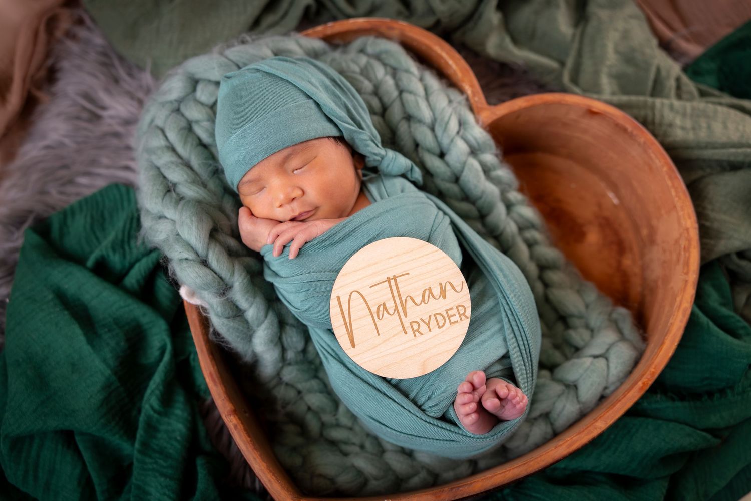 Sleeping newborn baby in a teal wrap and hat resting in a wooden bowl with a personalized name sign for a newborn photo session.