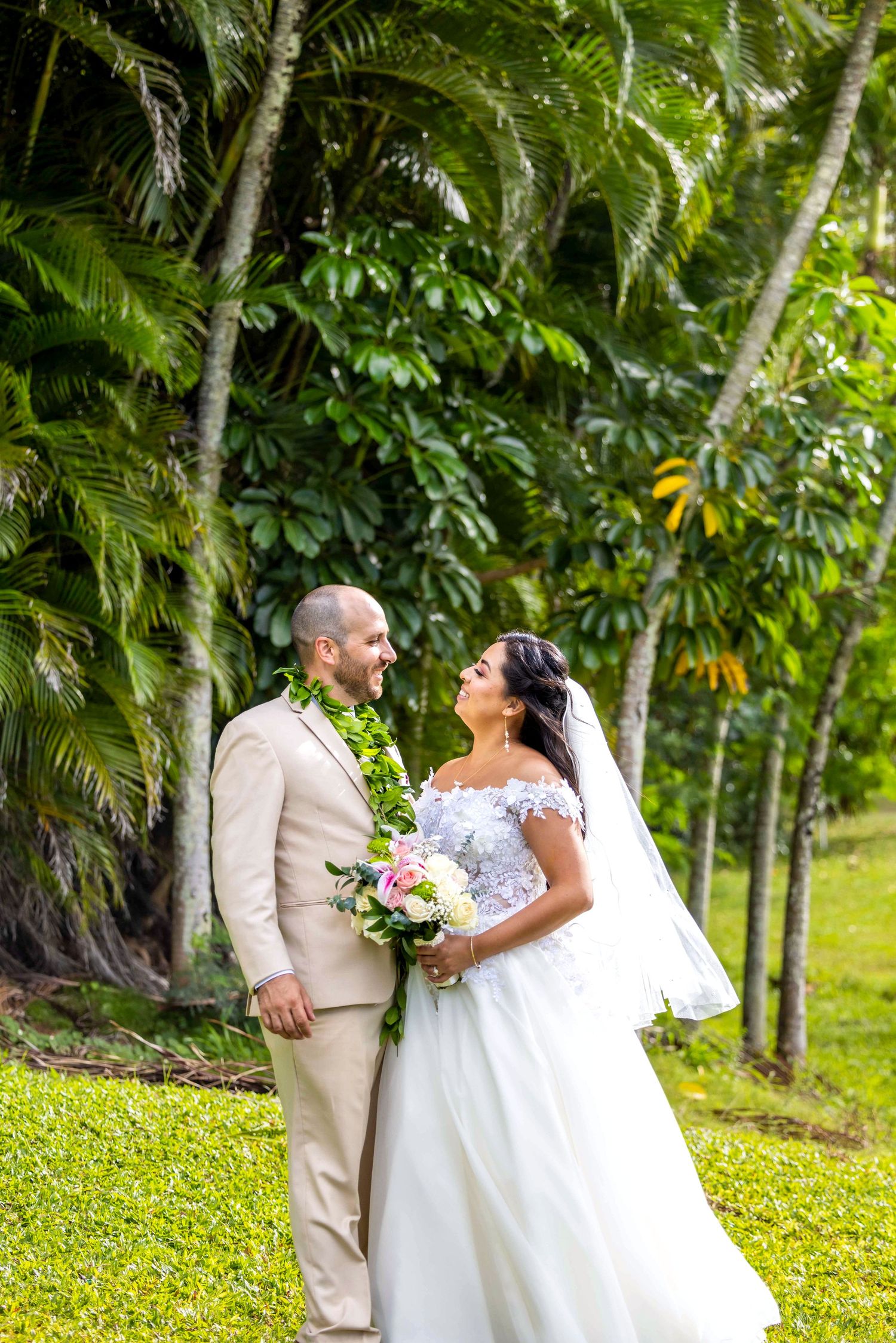 Bride in a white lace gown and veil and groom in a tan suit with a green lei share a tender moment among tropical palm trees. Lahaina
