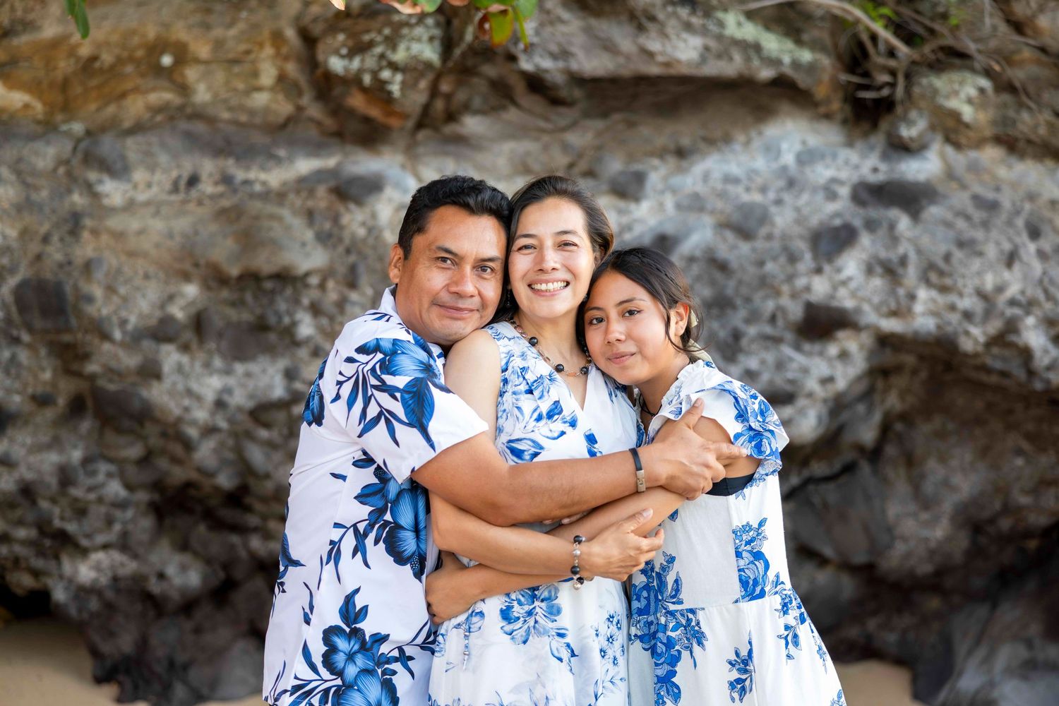 Man, woman and teenage girl in matching blue floral Hawaiian outfits share a warm hug in front of a rocky coastal cliff.