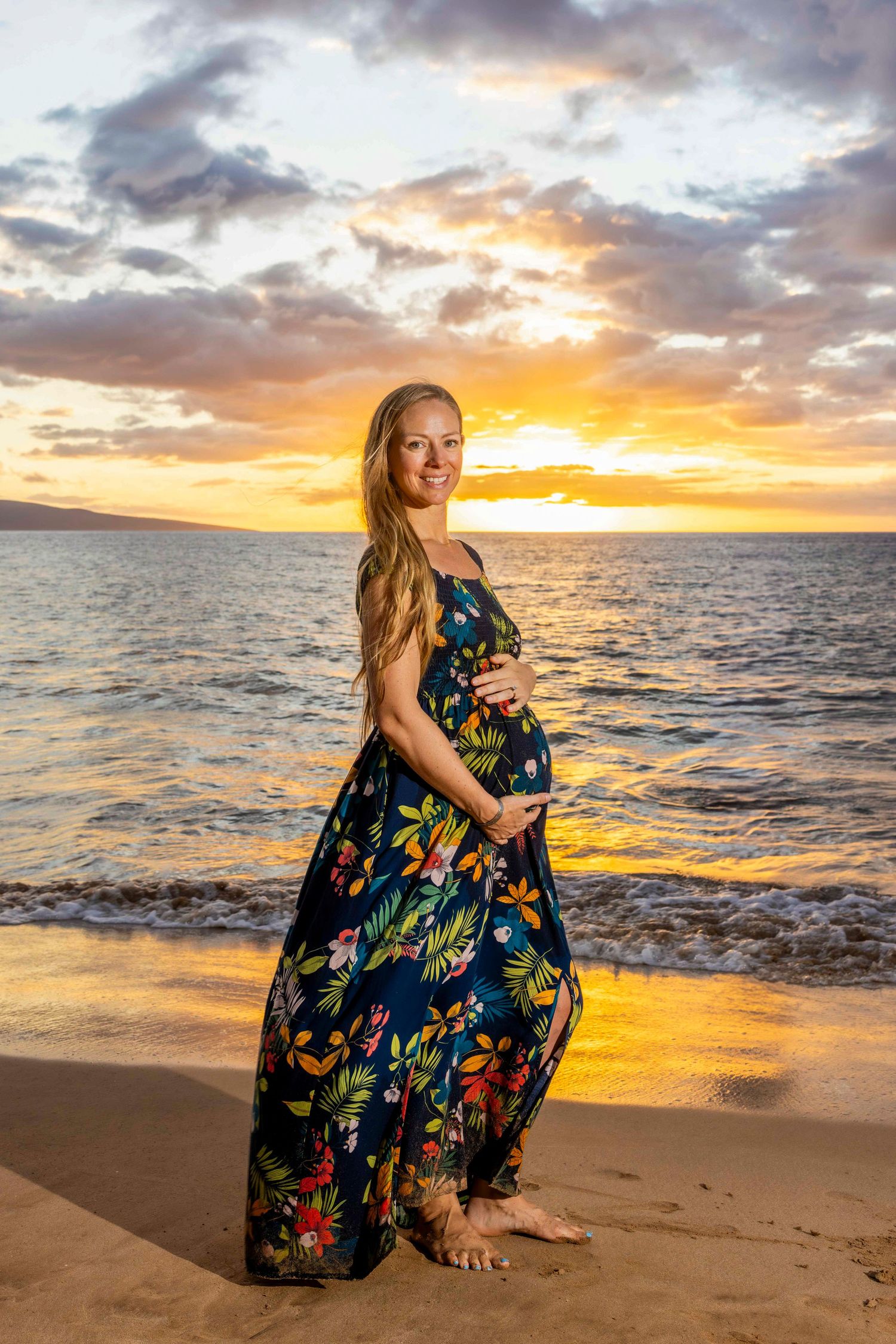 Pregnant woman in a colorful floral maxi dress cradles her baby bump on a sandy beach at golden sunset with ocean waves behind her. Wailea