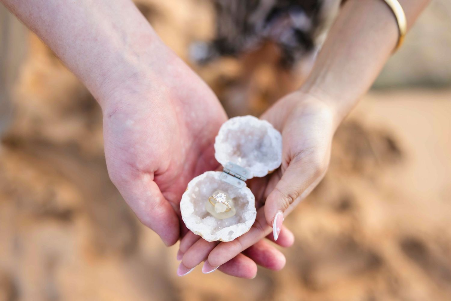 Two hands cupping an open pearl-lined shell that holds a small pearl, set against a soft, sandy beach background.