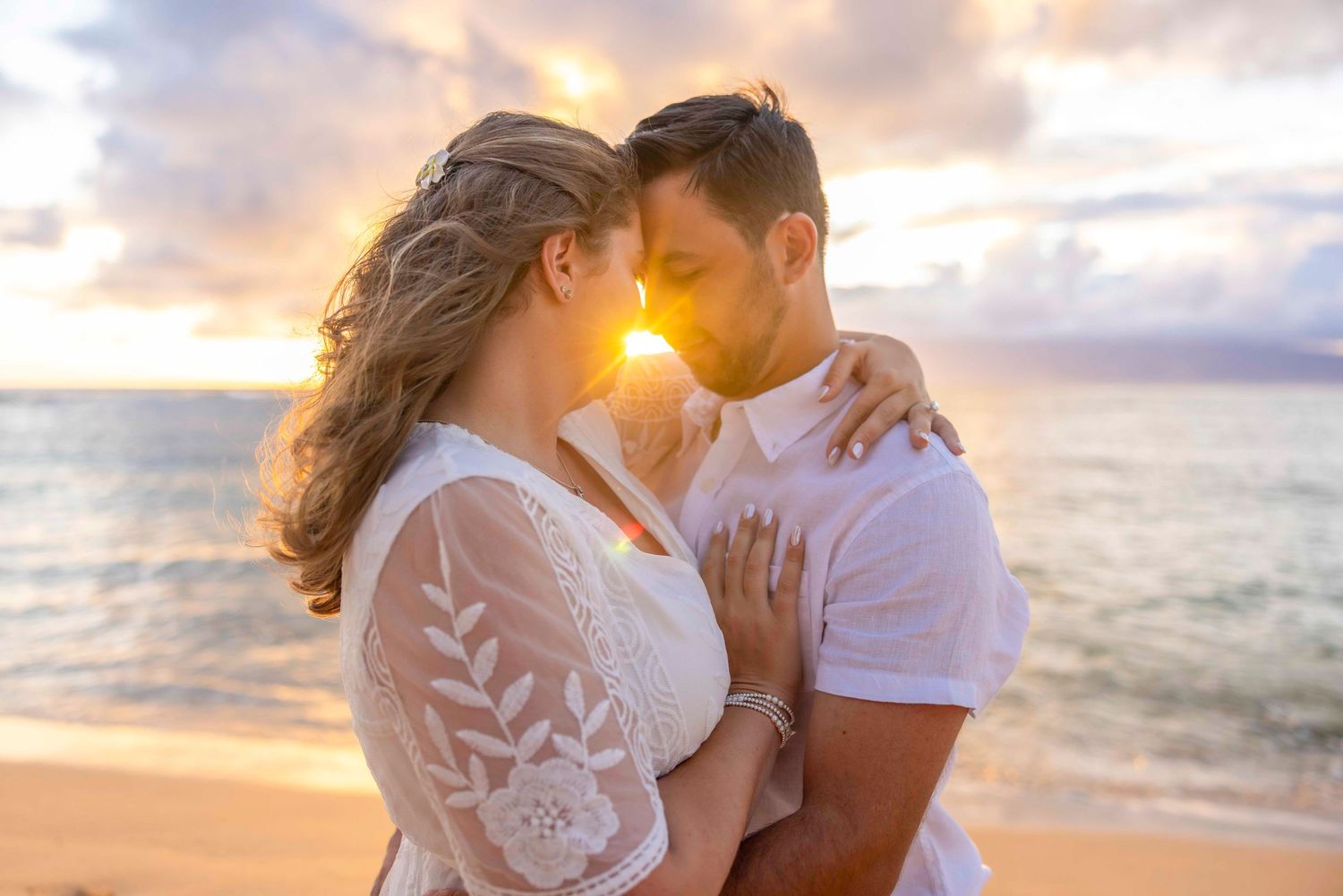 Romantic couple embracing foreheads together on a sandy beach at golden sunset with ocean waves and a glowing sky in the background. Kapalua
