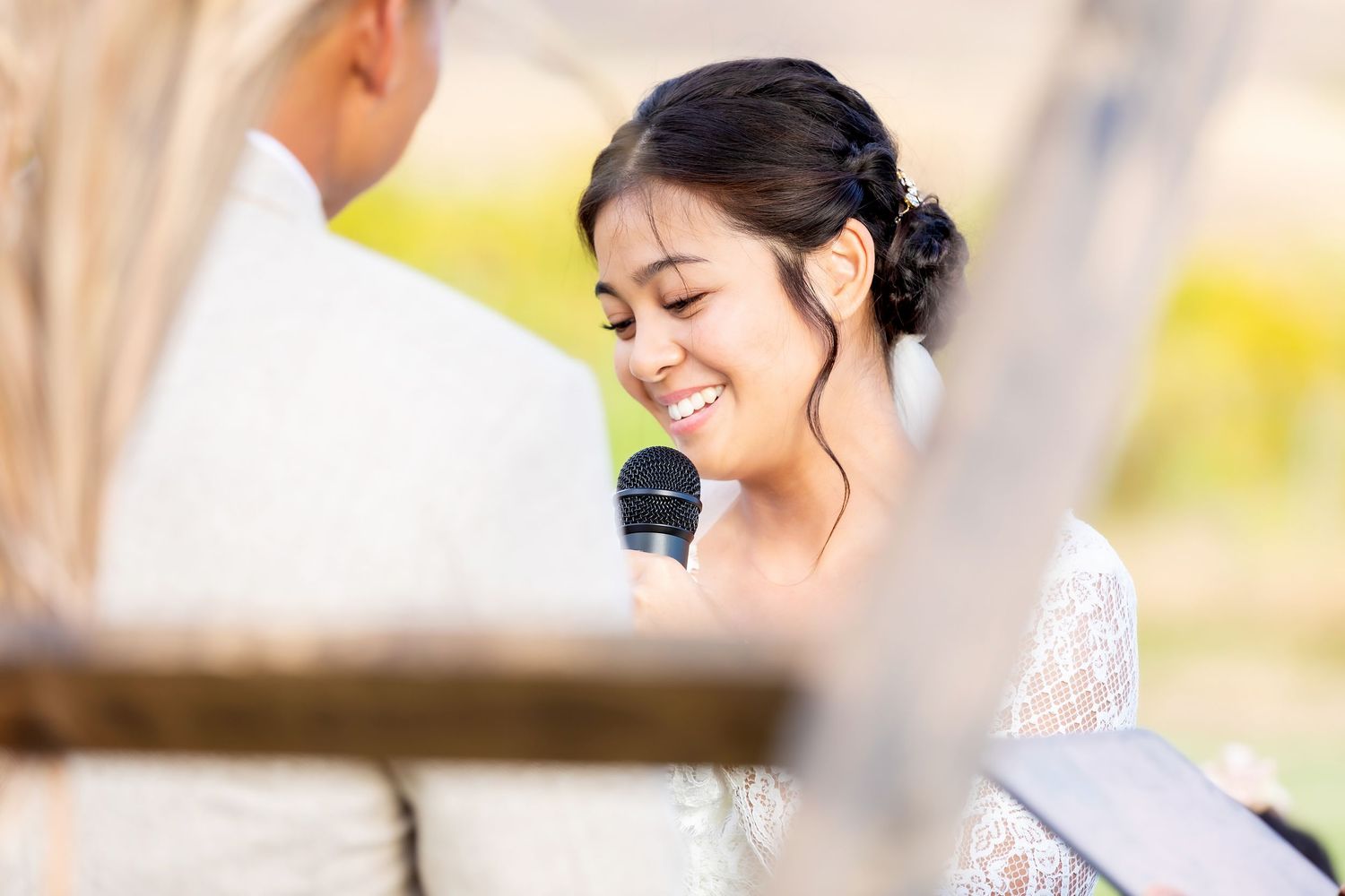 Smiling bride in a lace wedding dress with an elegant updo speaks into a microphone during an outdoor ceremony while facing her groom.