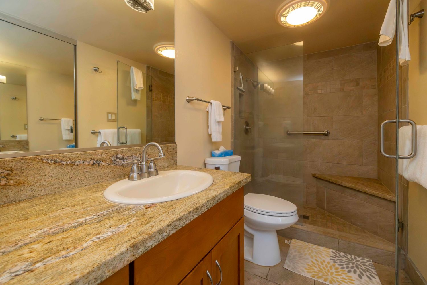 Bathroom with granite vanity, oval sink, wood cabinets, large mirror, toilet, and tiled walk-in glass shower with built-in bench.