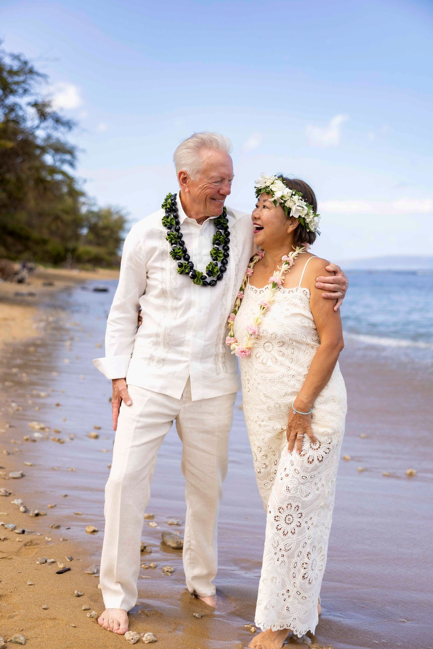 Smiling elderly couple in white linen outfits and floral leis embrace while walking barefoot along a sunny tropical beach by the ocean.