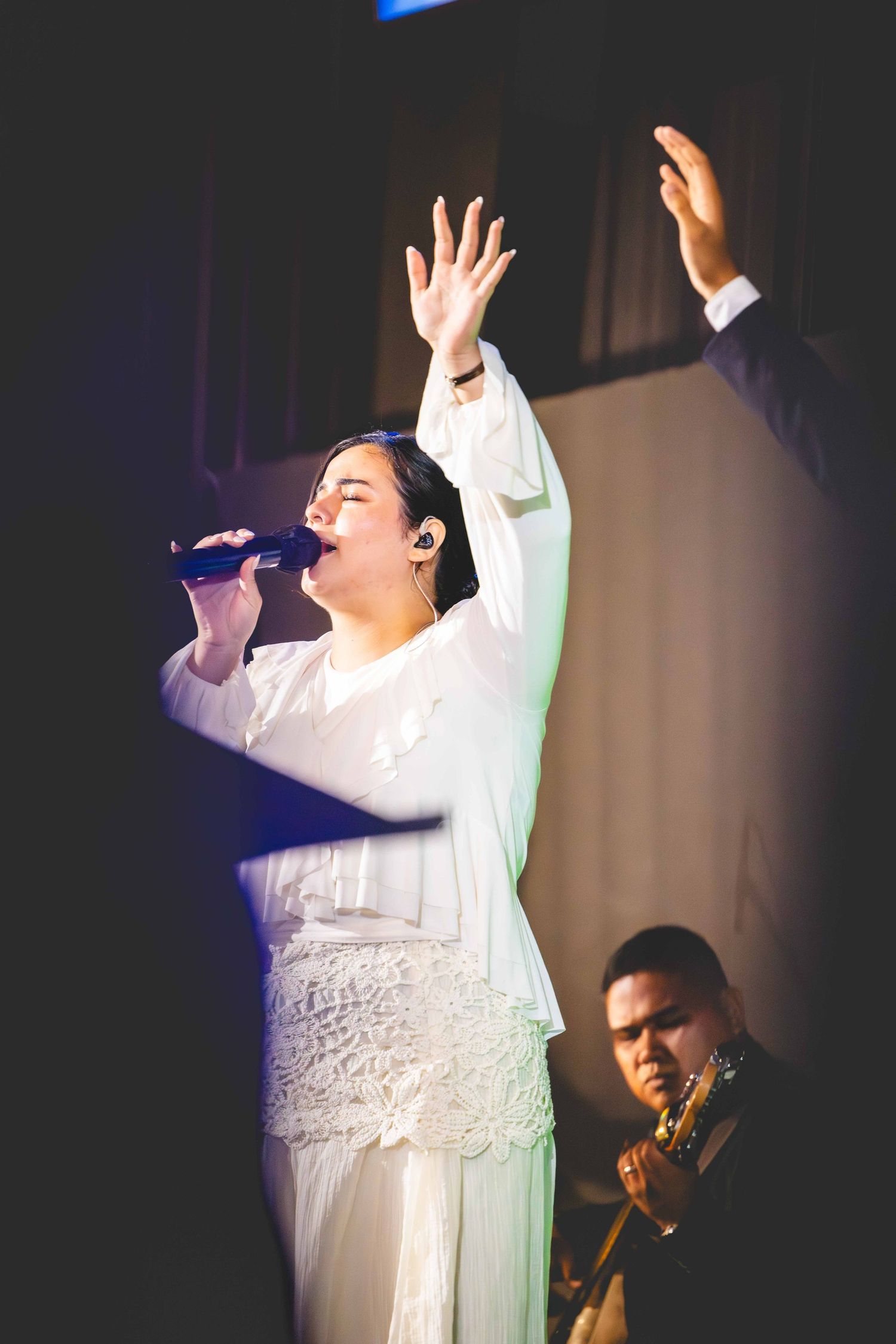 A woman in a white lace outfit sings into a microphone with raised hands while a guitarist plays behind her on a dimly lit stage.