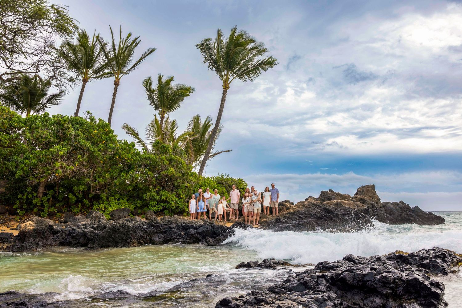 Group of people posing together on volcanic rocks beside crashing ocean waves, with palm trees and tropical greenery under a cloudy sky.