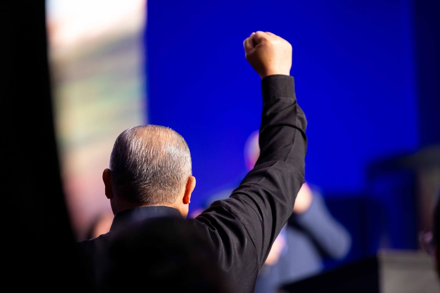 Man with gray hair in a dark jacket seen from behind raising a clenched fist in the air before a bright blue stage backdrop.
