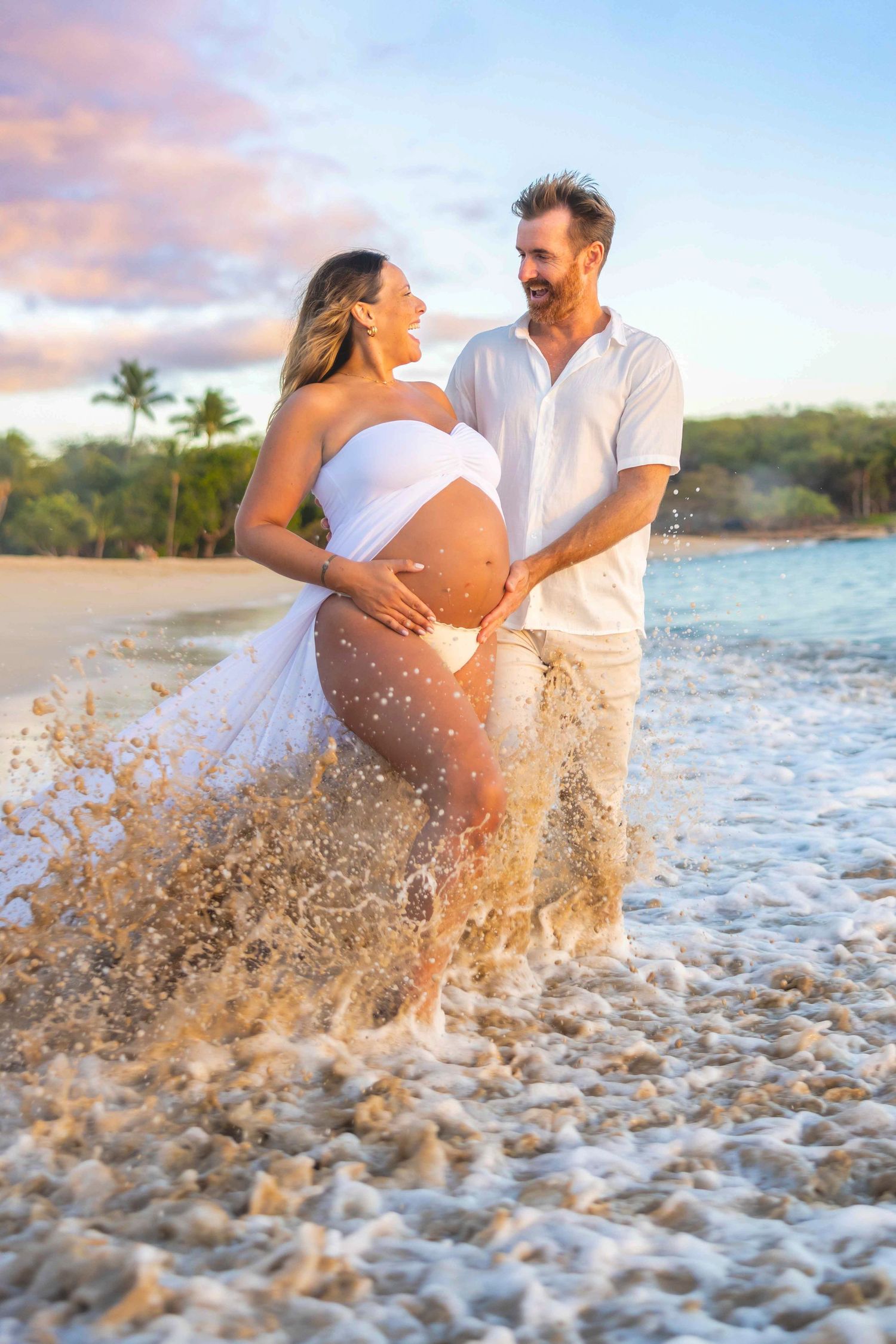 Pregnant woman in a white dress and her partner laughing together in splashing ocean waves on a tropical beach at sunset. Lana'i
