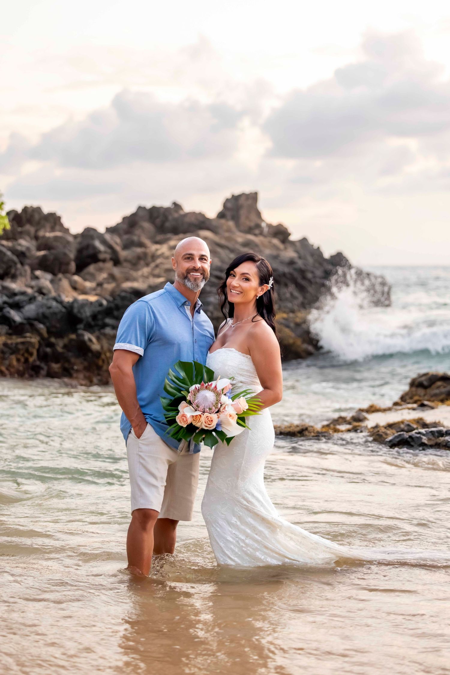 Bride in a white gown and groom in a blue shirt stand in shallow water holding a tropical bouquet as waves crash on rocks behind them.