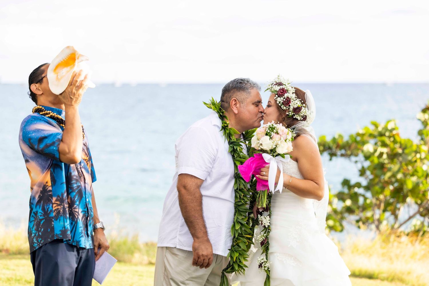 Couple in leis share a kiss at a seaside Hawaiian wedding ceremony as an officiant blows a conch shell nearby. Lahiana