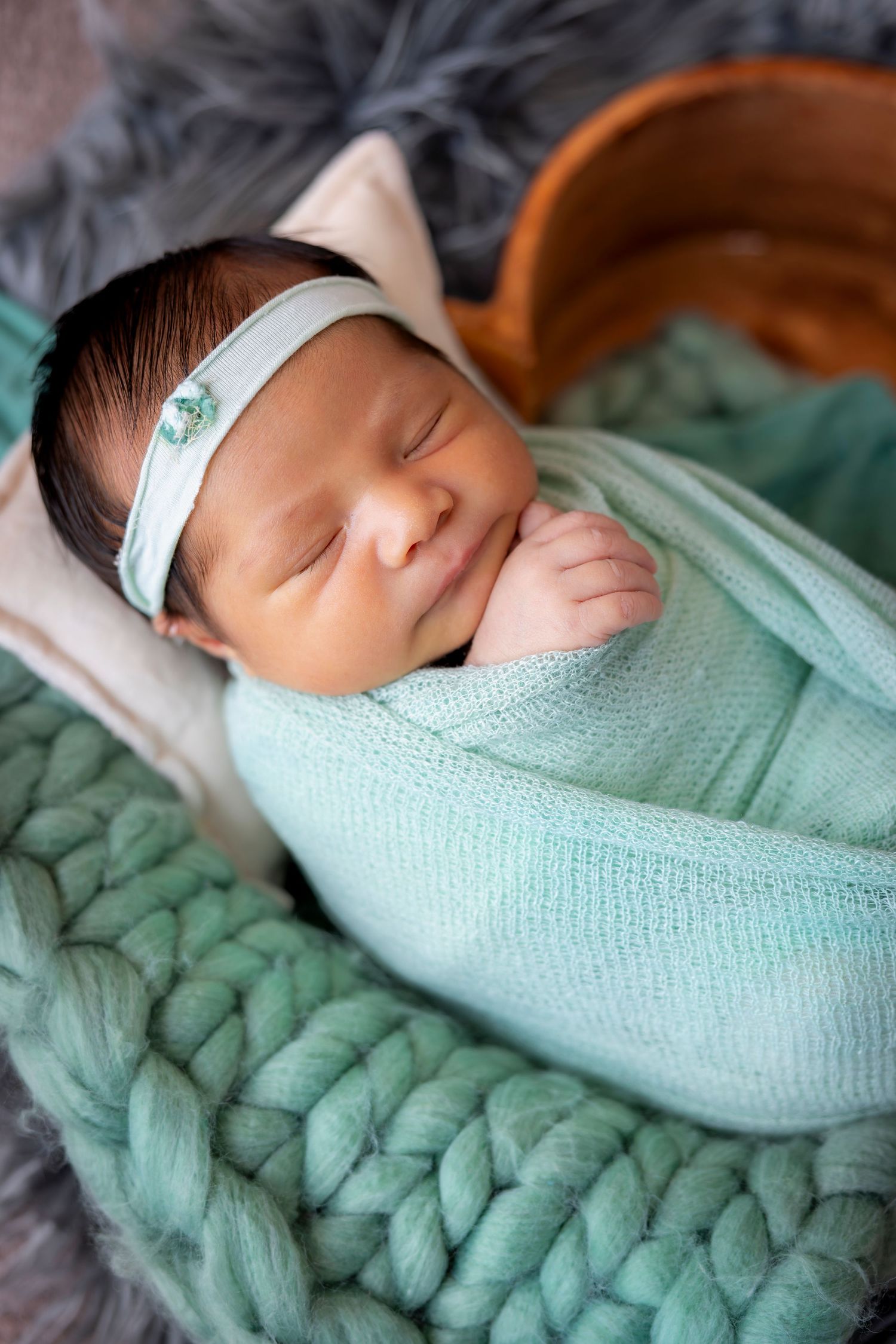 Sleeping newborn girl in a mint green swaddle and headband rests in a basket on a chunky knit blanket.