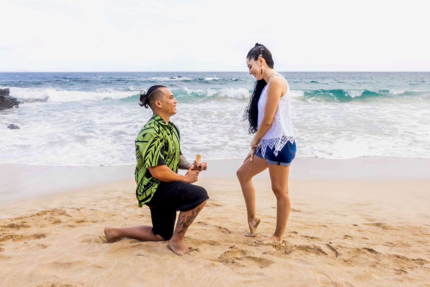 Man in a green floral shirt kneels on one knee on a sandy beach proposing to a smiling woman with ocean waves behind them. Ironwood