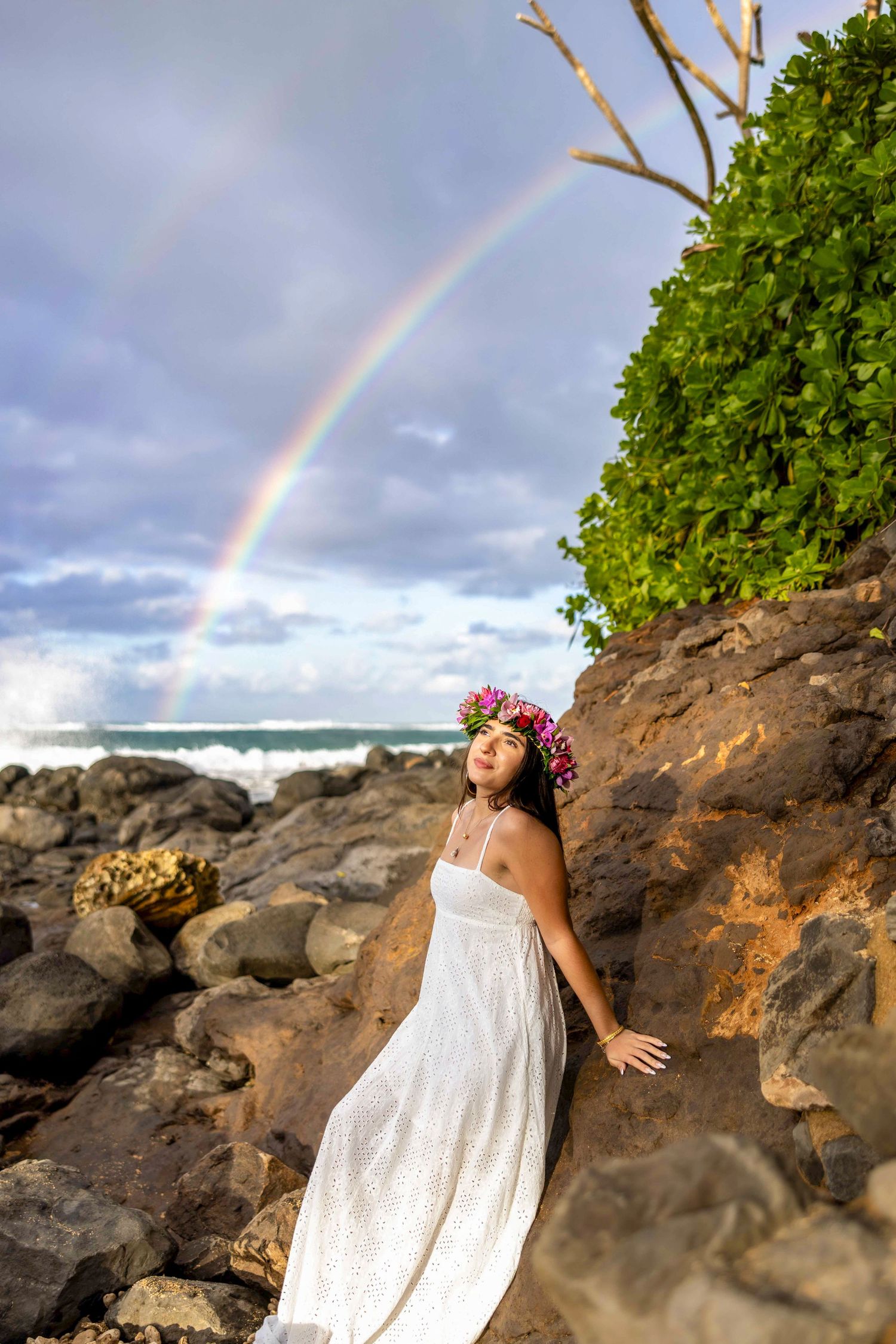 Woman in a white dress and flower crown leans against volcanic rocks on a coastline beneath a vivid double rainbow. Paia