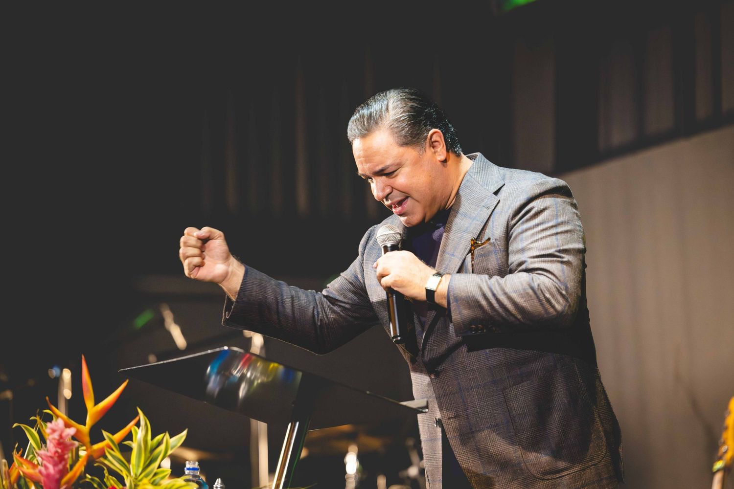 A man in a gray blazer sings passionately into a microphone with a raised fist on stage beside colorful tropical flowers.