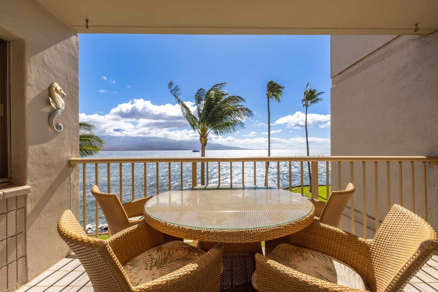 Oceanfront balcony with a wicker dining set, glass table, and wooden railing overlooking palm trees, blue water, and distant island views.