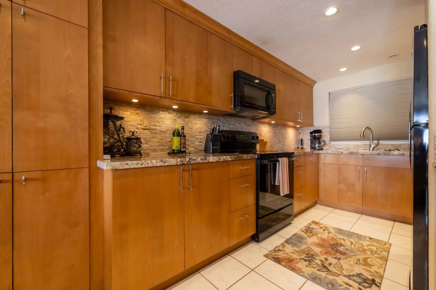 Bright kitchen with light wood cabinetry, granite countertops, black range and microwave, and a decorative rug on the tile floor.