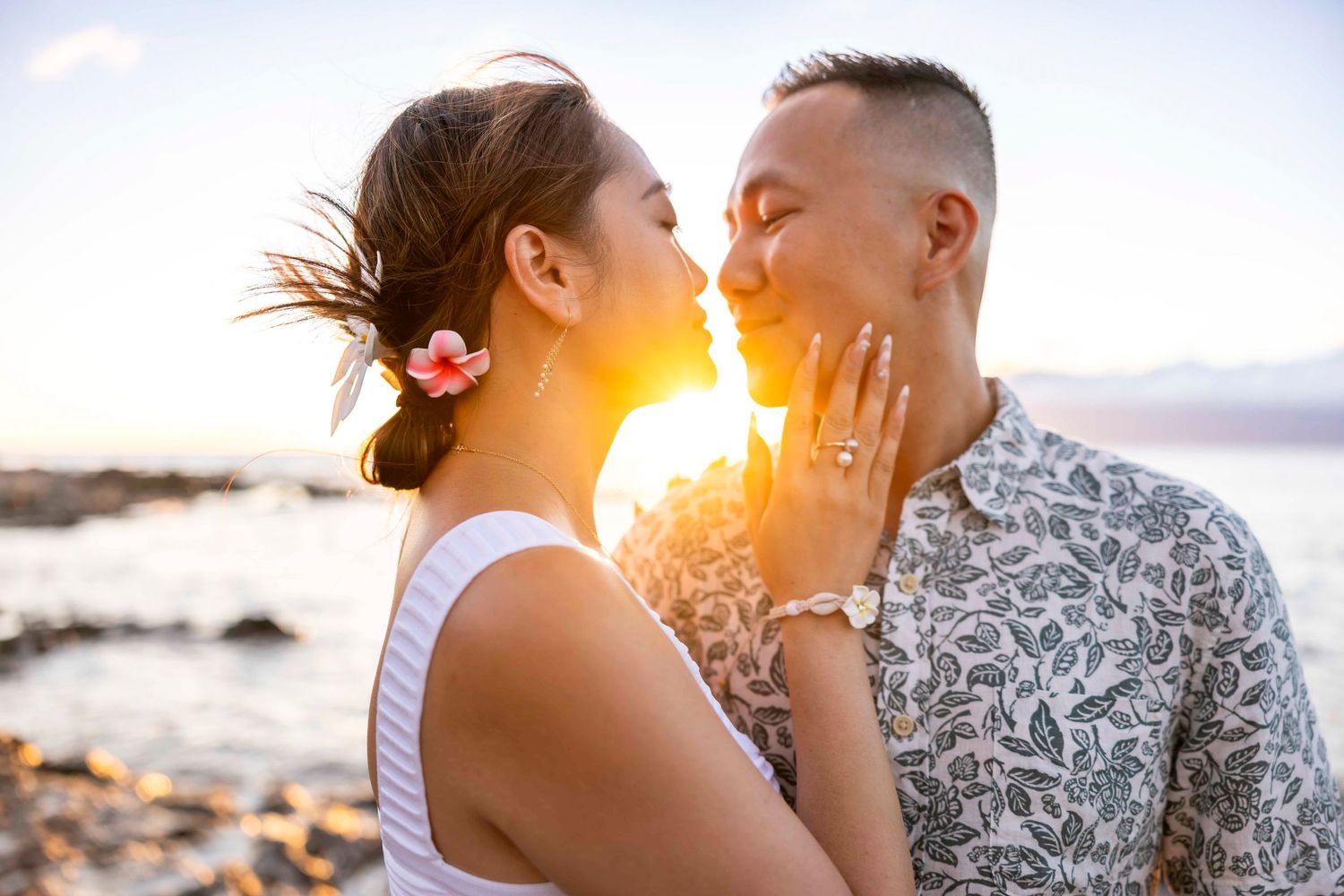 Couple embracing and almost kissing at sunset on a rocky beach, she wears a pink flower in her hair and a ring, he wears a floral shirt.
