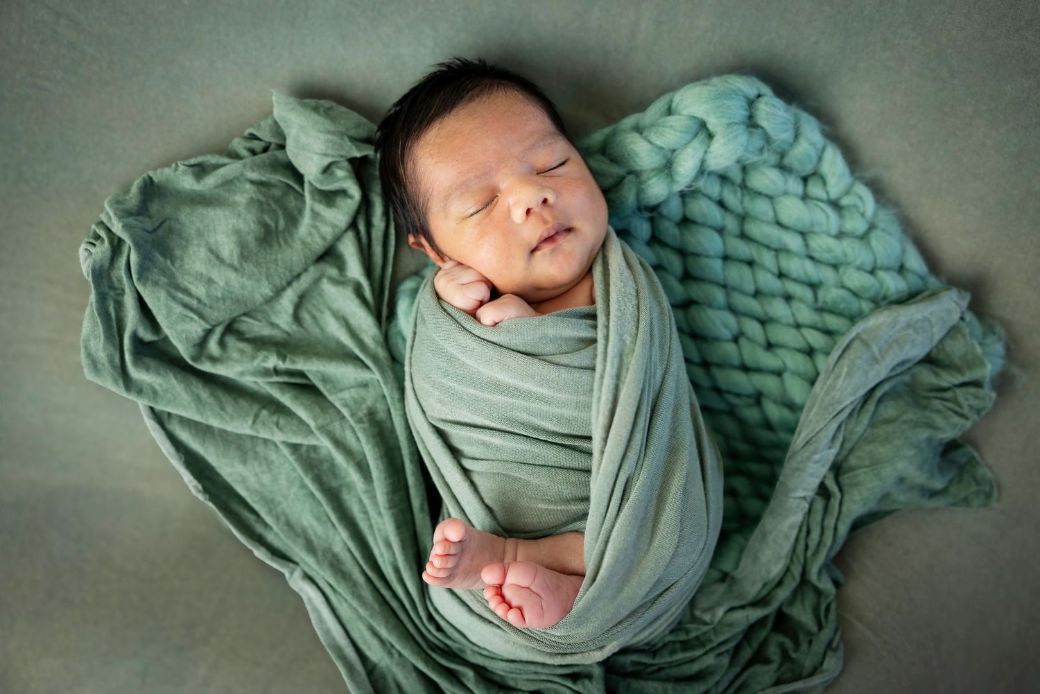 Peaceful newborn swaddled in sage green fabric rests on a heart-shaped blanket with chunky knit texture.