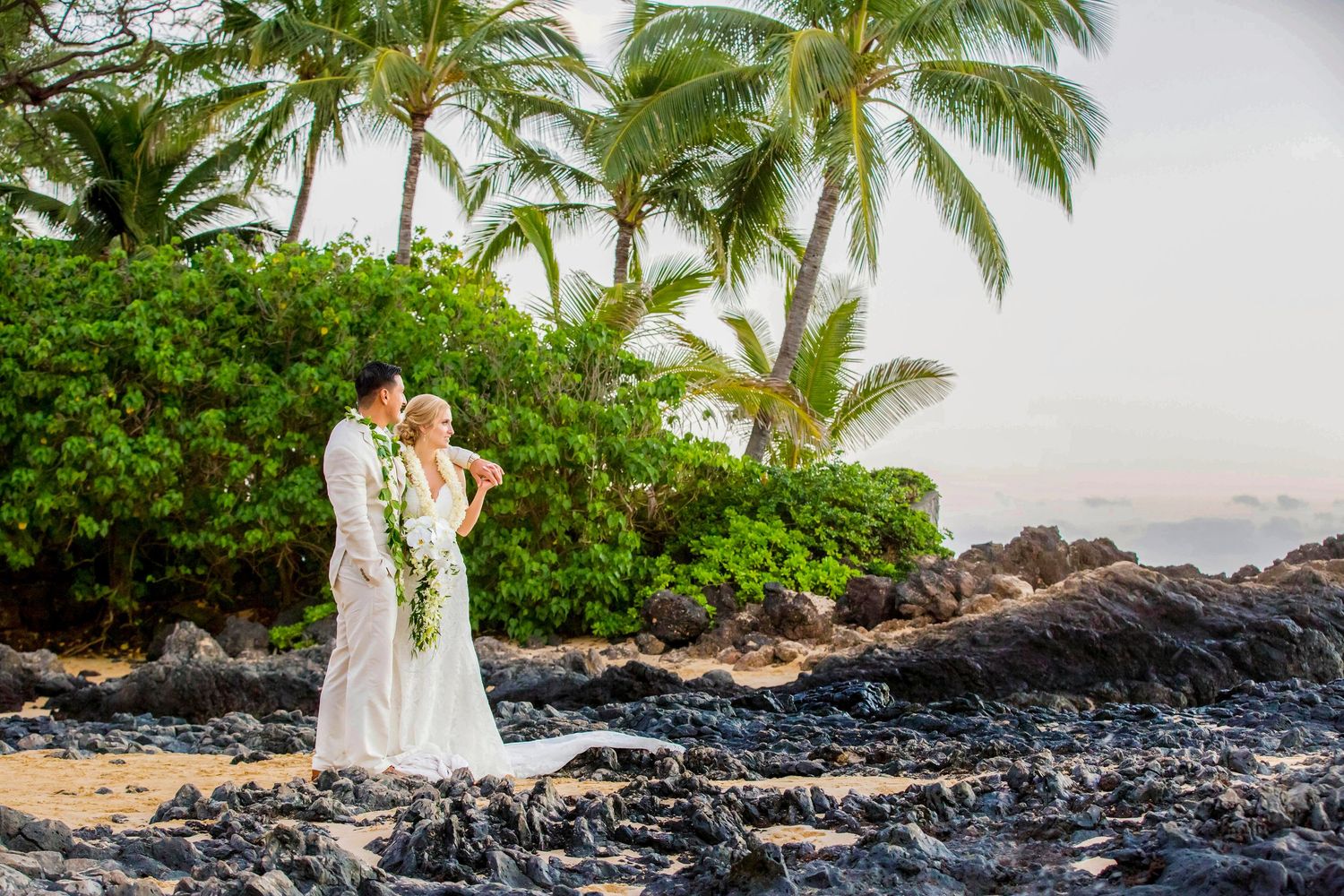 Bride and groom in white wedding attire embrace on a rocky tropical beach framed by palm trees, lush greenery, and a soft sunset sky. Makena