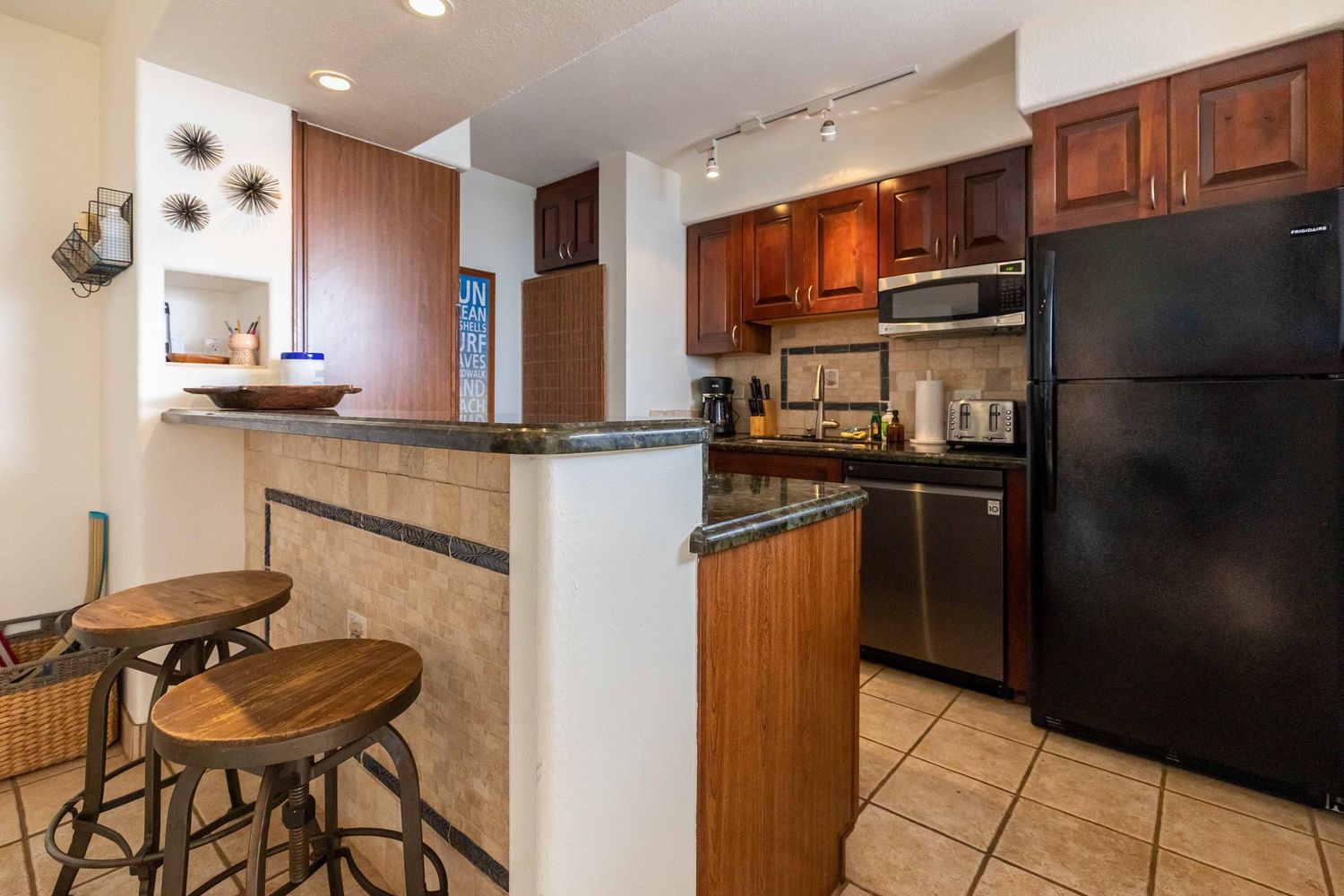 Condo kitchen with dark wood cabinets, black refrigerator, tile floor, breakfast bar and two wooden stools.