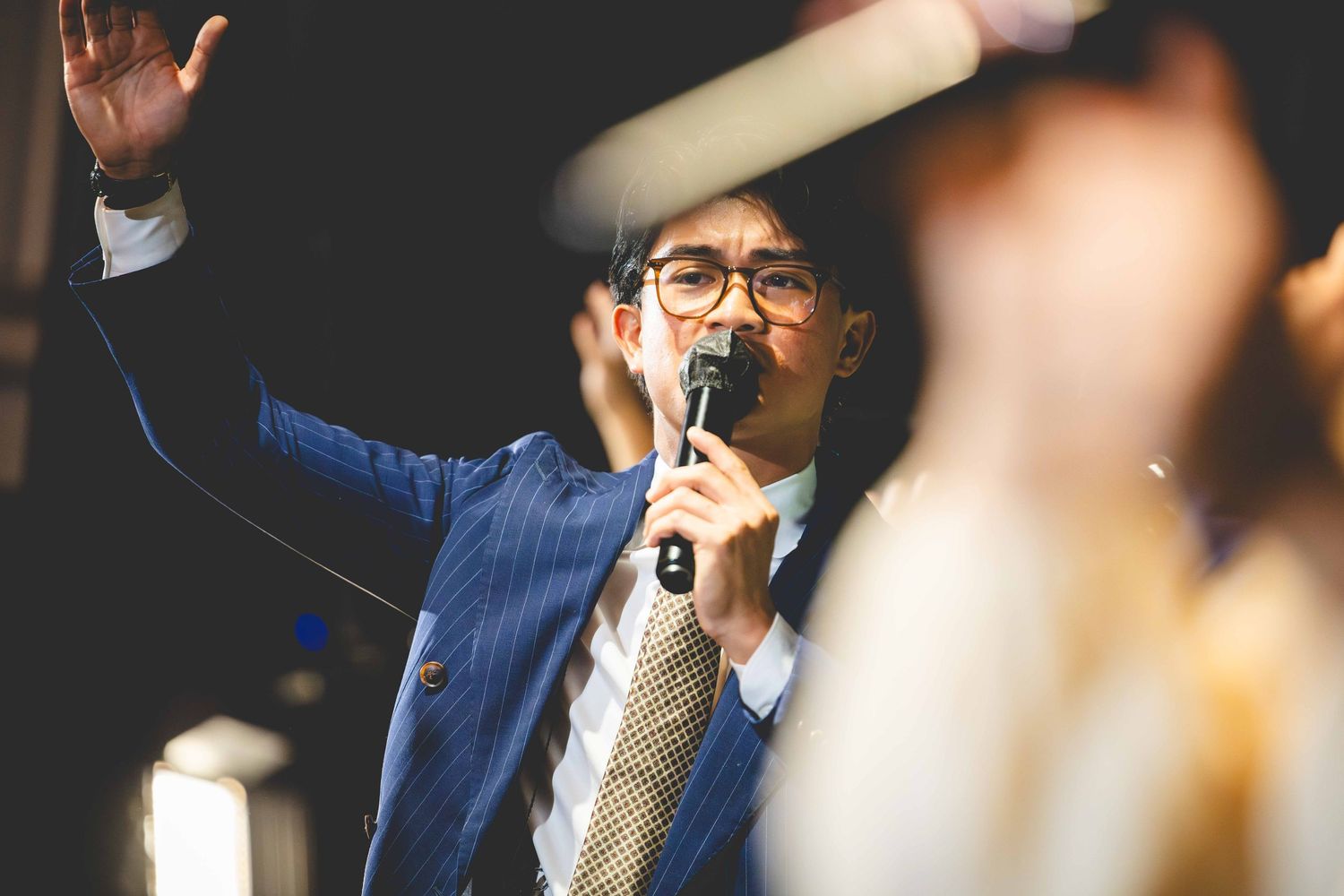 A man in glasses and a blue blazer speaks into a microphone with one hand raised while addressing a blurred crowd.