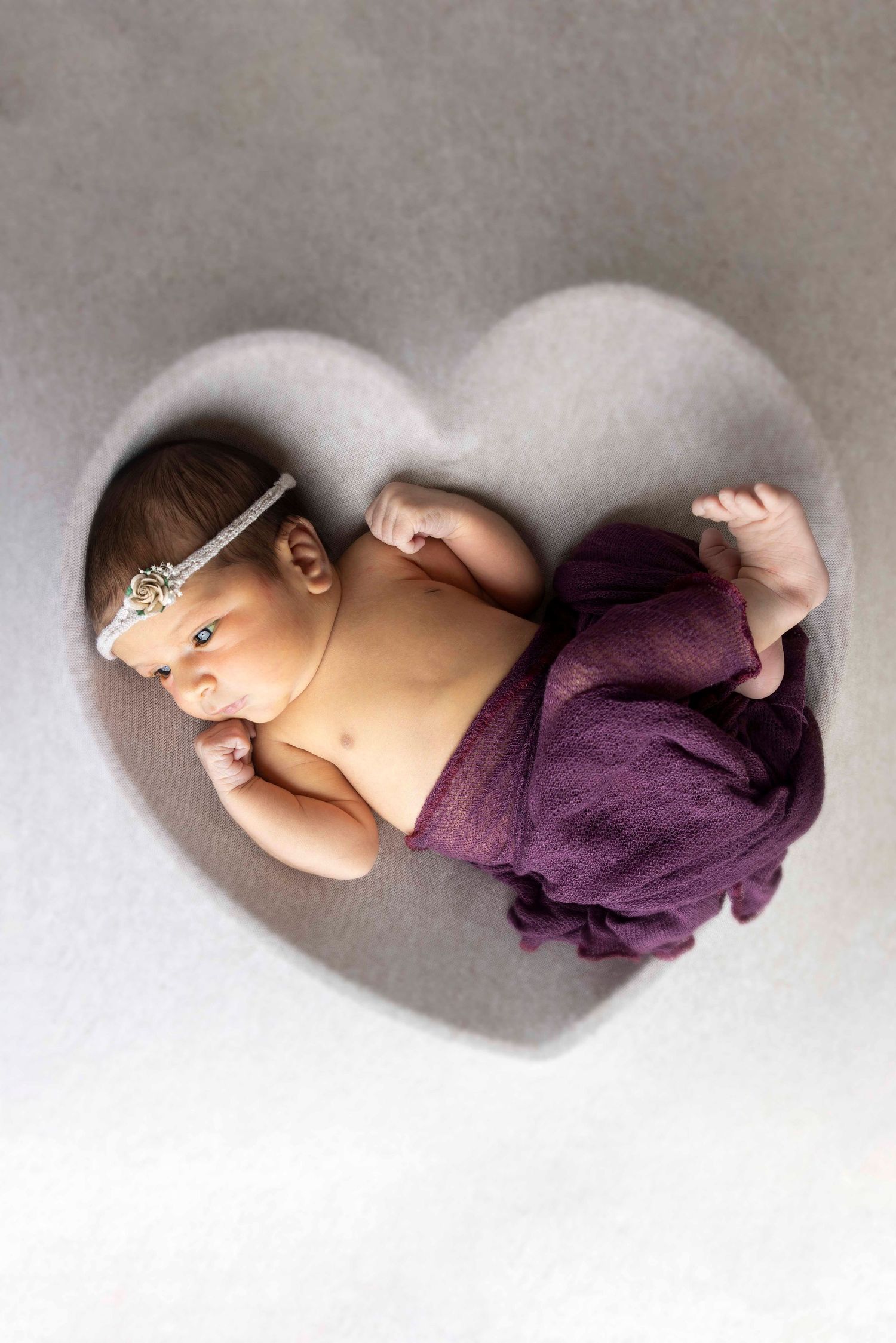 Newborn baby girl with a jeweled headband lies in a heart-shaped nook wrapped in a purple cloth.