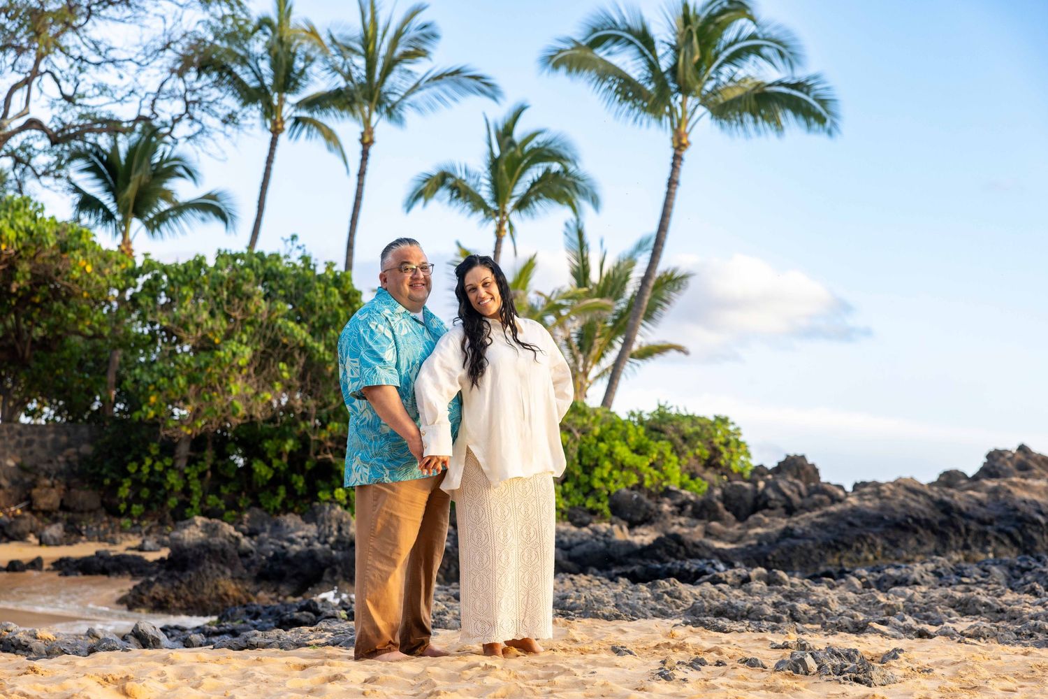 Couple in coordinated island attire embracing on a sandy tropical beach with palm trees, lava rocks, and blue sky in the background.