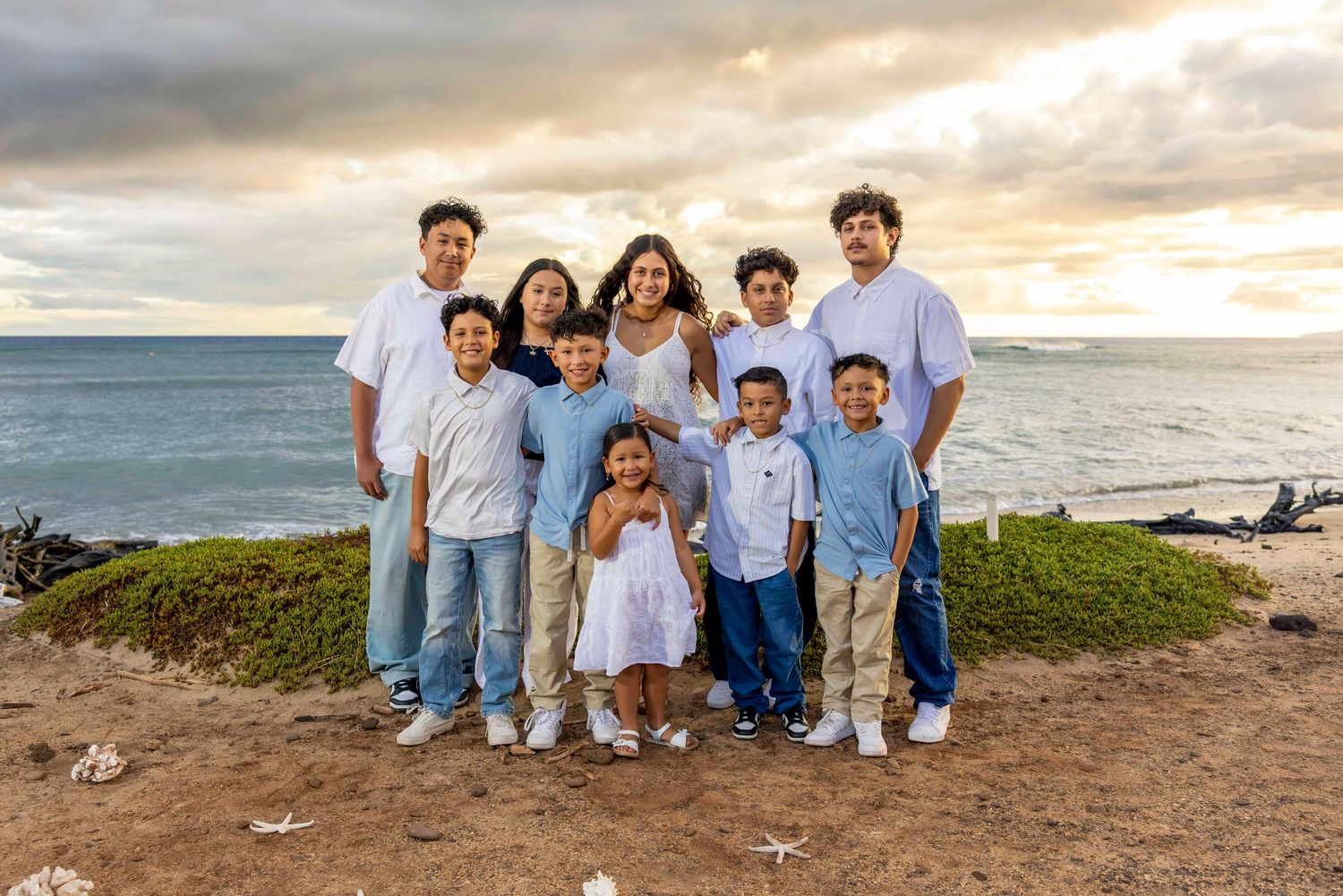 Large family in white and light blue outfits poses on a grassy bluff overlooking the ocean at golden hour with children in front.