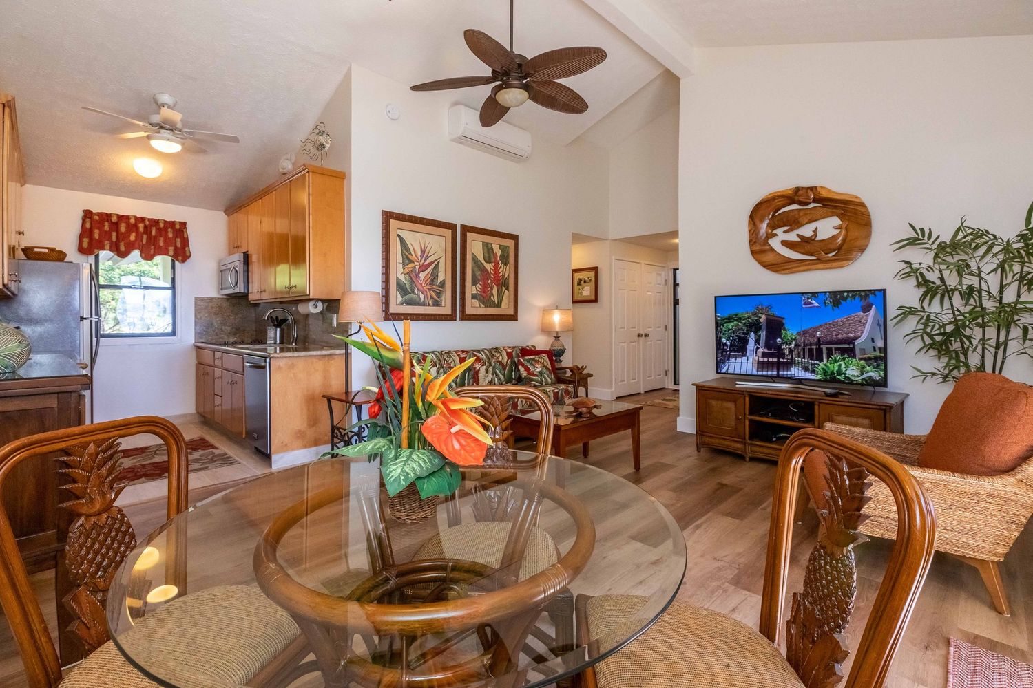 Bright open-concept vacation rental living area with vaulted ceiling, tropical flower centerpiece, glass dining table, TV, and kitchen.