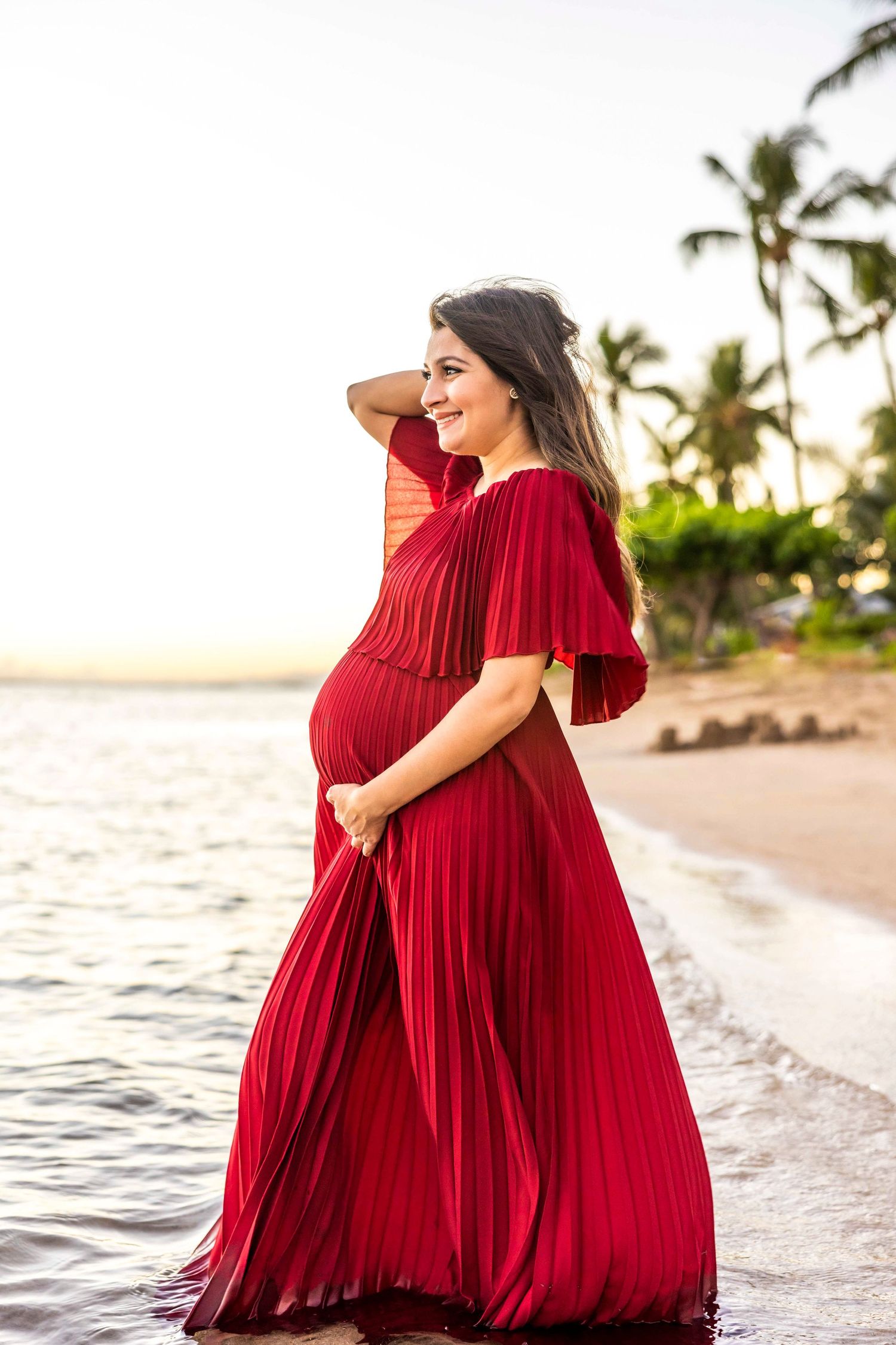 Pregnant woman in a flowing red pleated maternity gown standing in shallow water on a tropical beach with palm trees at sunset. Lahaina