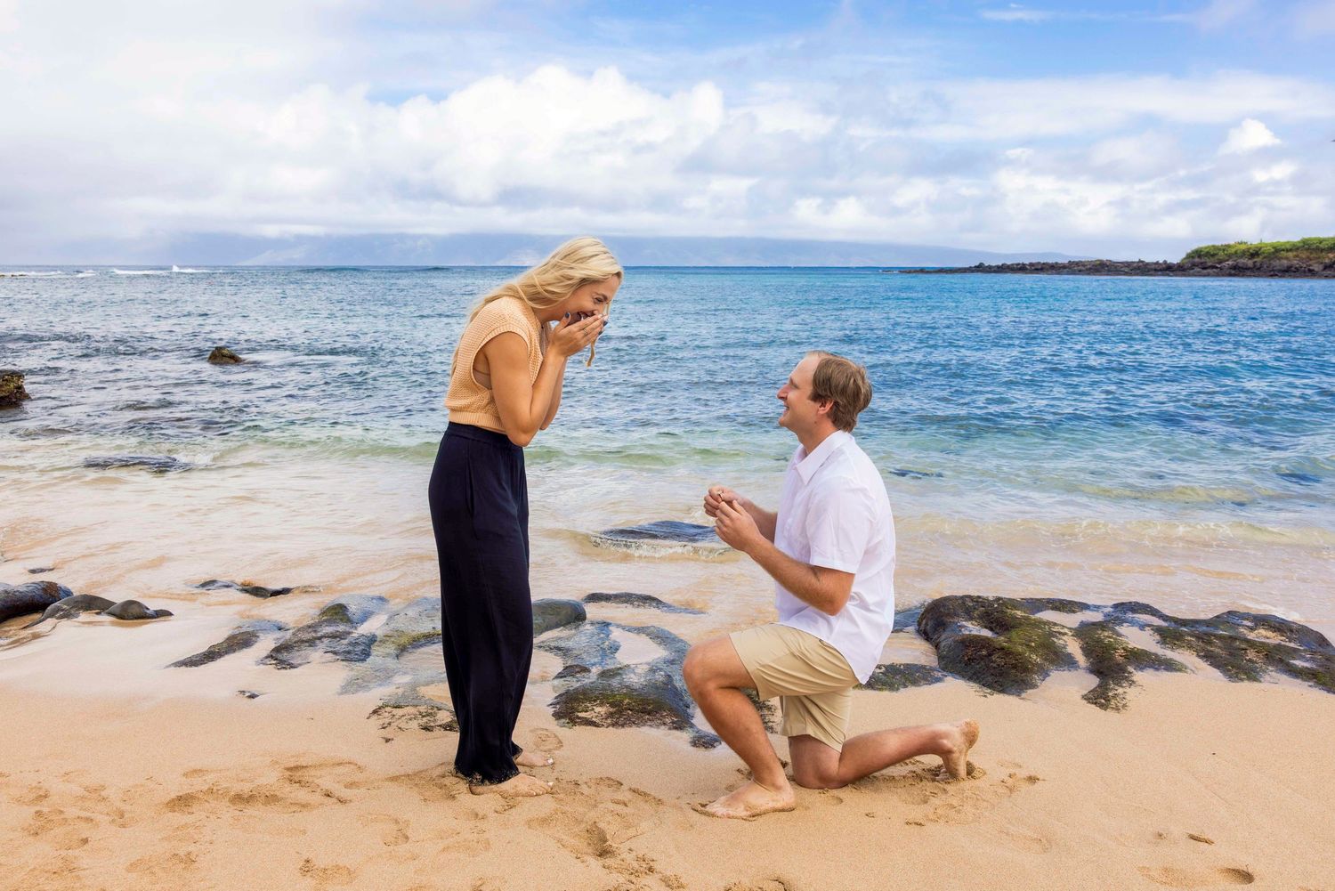 Man kneeling on one knee proposing to a surprised woman covering her mouth on a sandy tropical beach with turquoise ocean and rocks. Kapulua