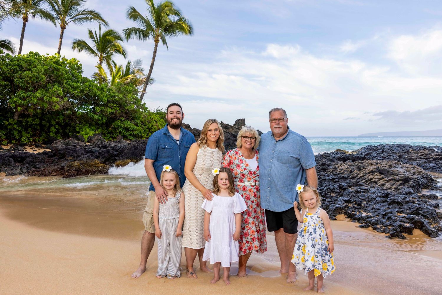 Family of seven with three young girls in white dresses pose together on a golden sand beach beside dark lava rocks and palm trees.