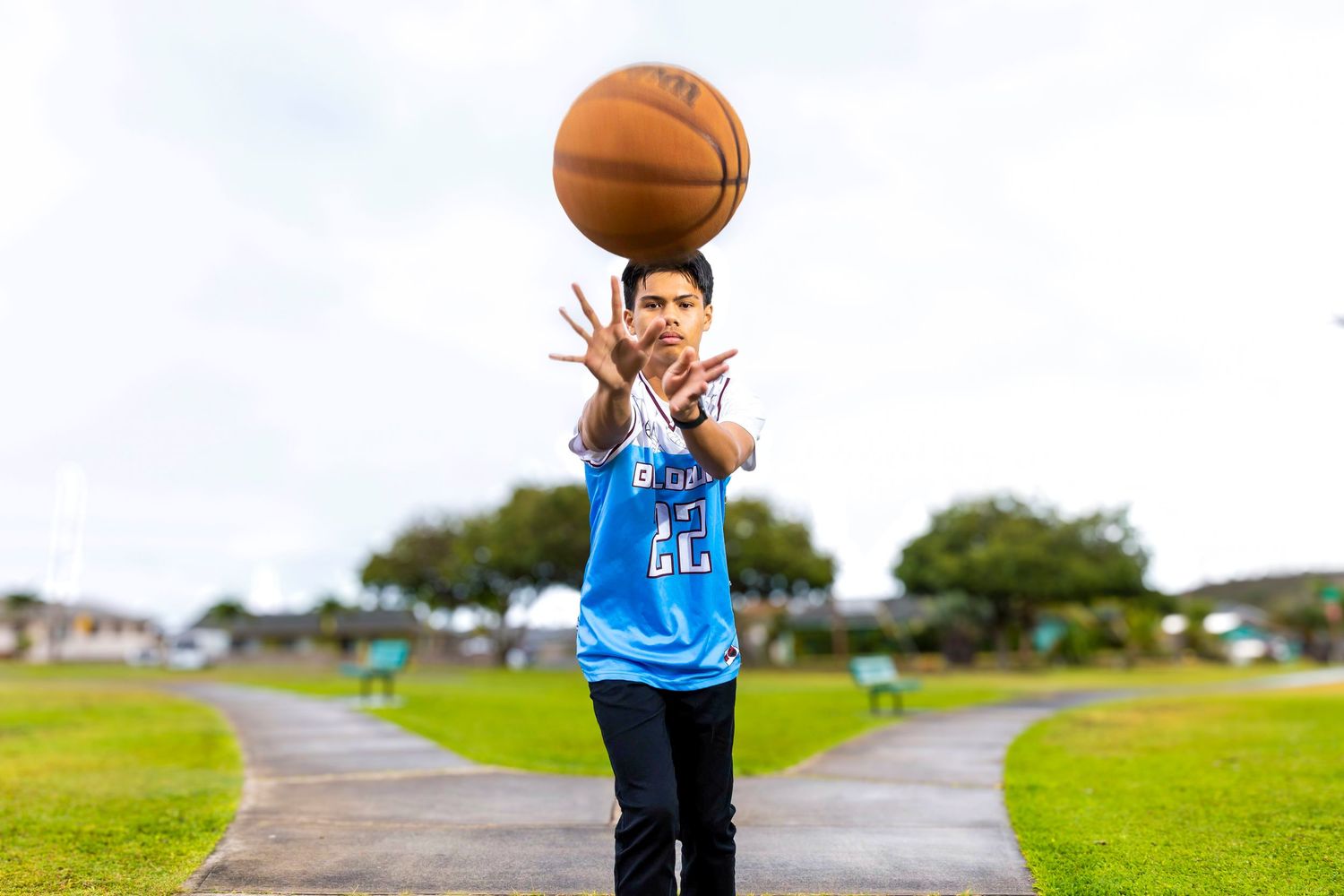 Young person in a blue basketball jersey tosses a basketball into the air on a paved path in a green park under a cloudy sky.