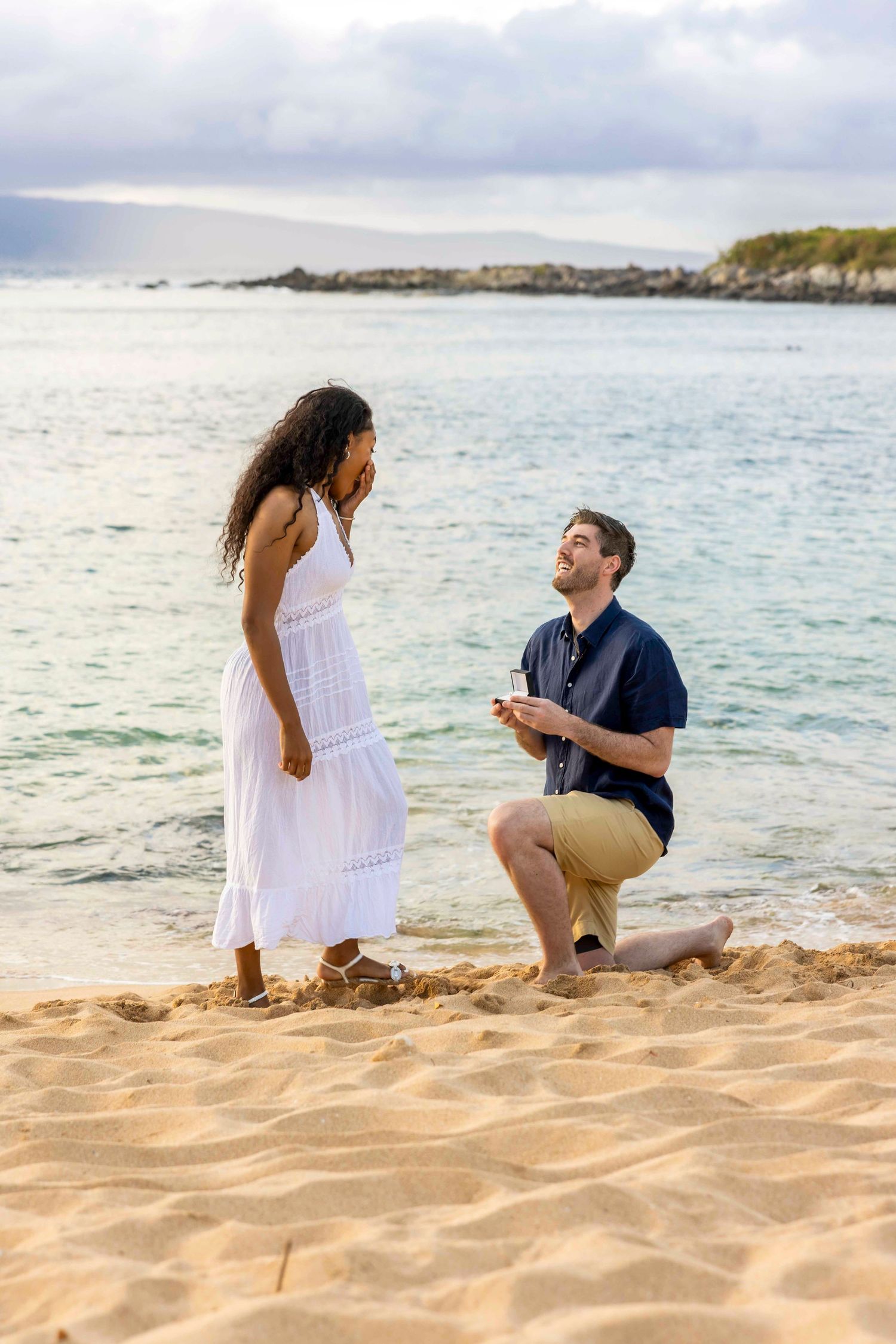Man kneeling on a sandy beach holds out a ring as a woman in a white dress covers her face in surprise during a seaside proposal. Kapalua