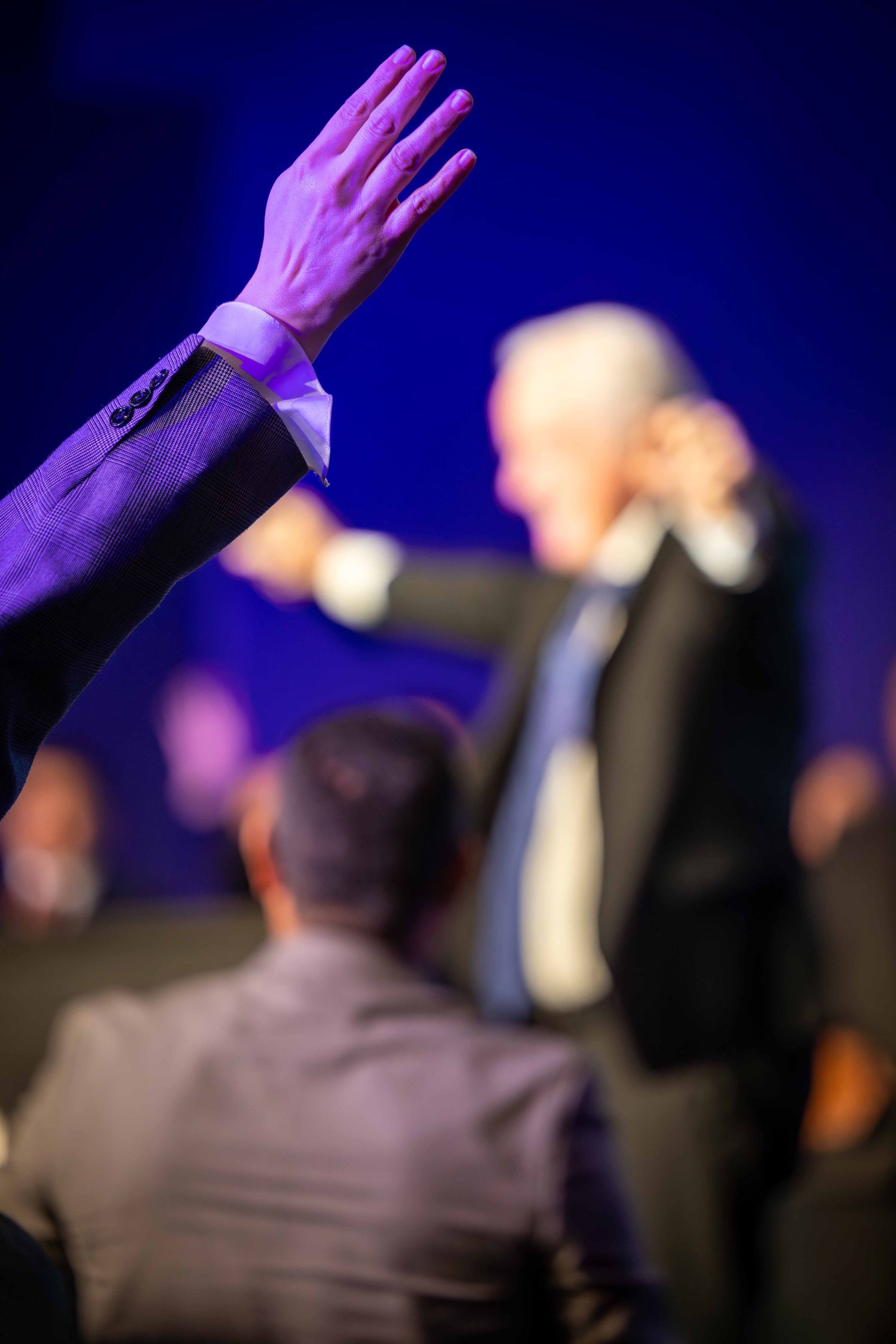 Raised hand in a suit sleeve in the foreground as a blurred older man in a dark suit gestures on stage before a seated audience.