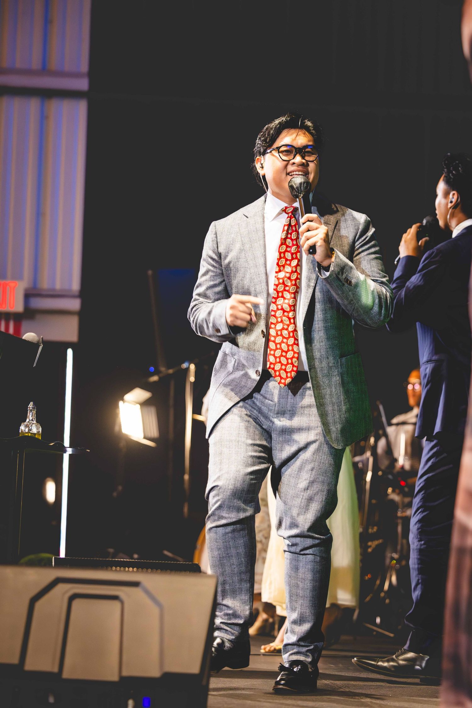Man in a gray plaid suit, glasses and red patterned tie speaks into a microphone while standing on a stage with another person nearby.