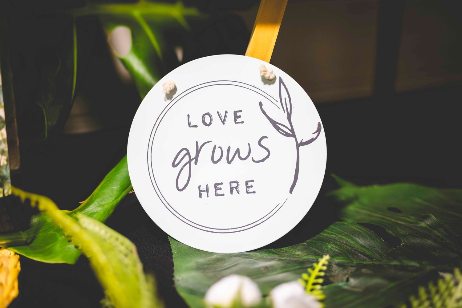 Round white sign reading Love Grows Here with a leaf branch design, hanging among lush green tropical leaves and white flowers.