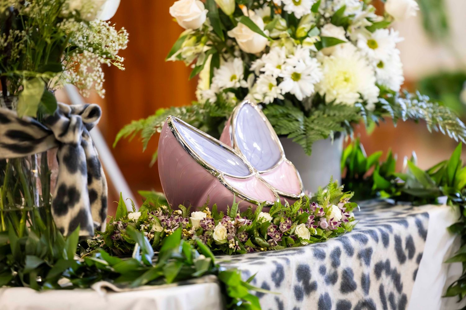 Pink satin ballet flats rest on a leopard print cloth among white flowers and green foliage in a decorative floral arrangement.