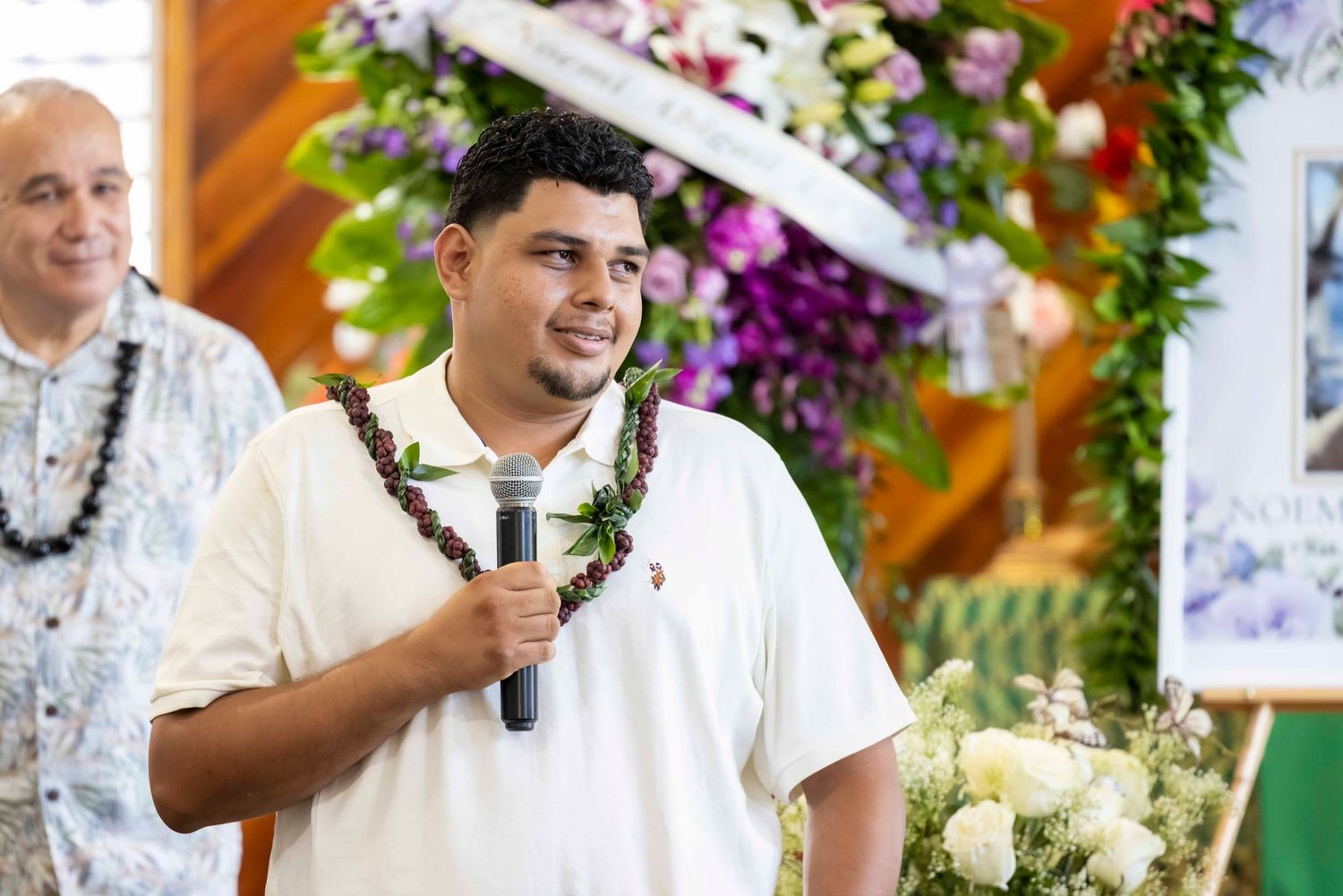 Smiling man in a white polo and leaf lei holds a microphone while speaking beside colorful floral arrangements at a ceremony.