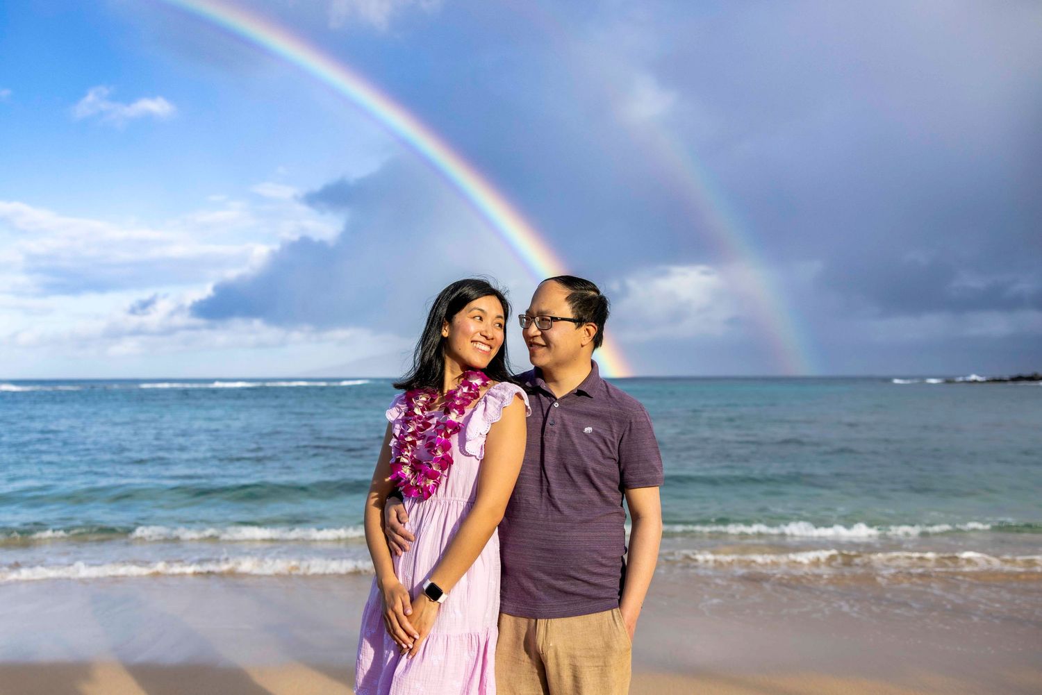 Smiling couple in matching purple outfits pose on a sandy beach with a vivid double rainbow arcing over the ocean behind them.