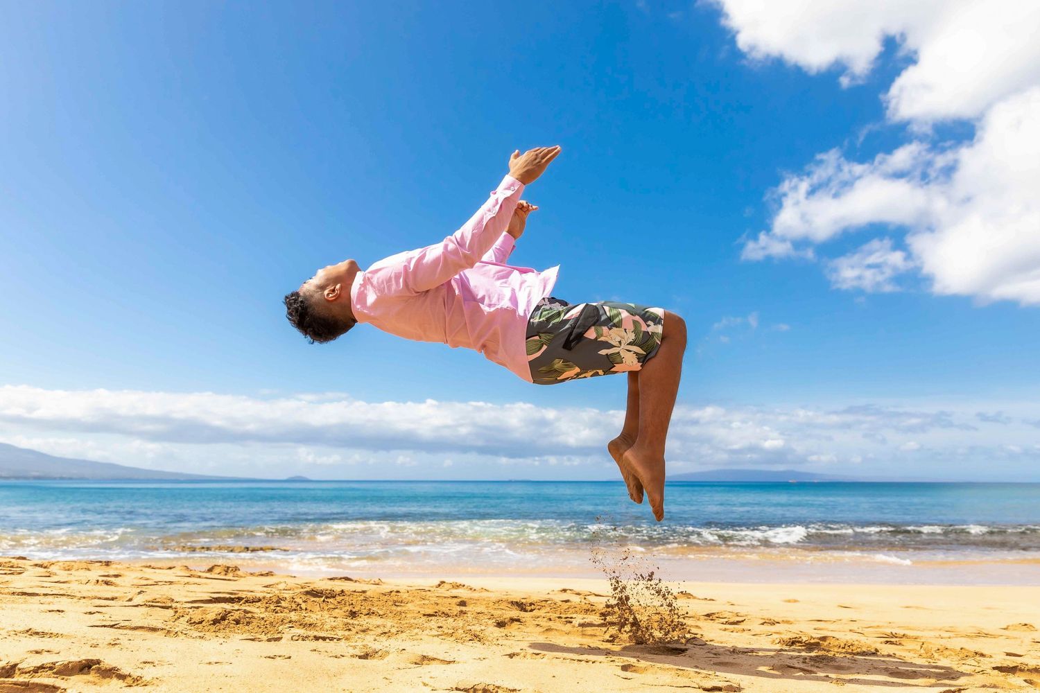 Person in a pink shirt and patterned shorts performs a mid-air backflip on a sunny sandy beach with turquoise ocean and blue sky. Kealia