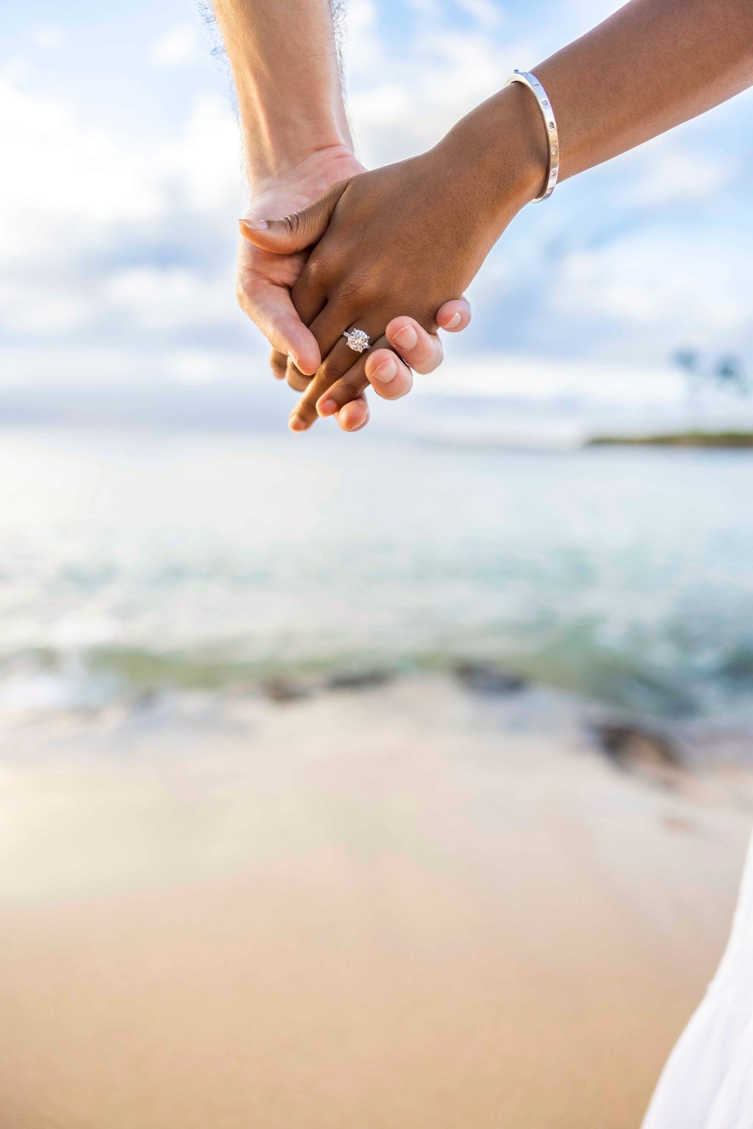 Couple holding hands on a sandy beach at sunset, with a sparkling diamond engagement ring on her finger and calm ocean waves behind. Kapalua