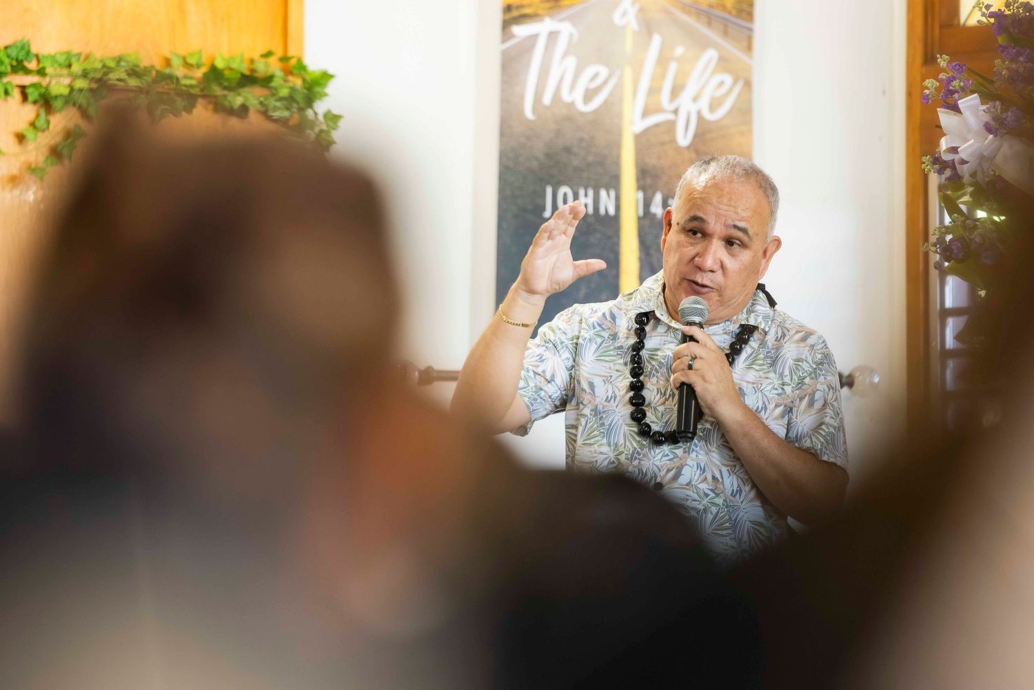 Man in a floral shirt and beaded necklace gestures while speaking into a microphone at a church service with a Bible verse sign behind him.