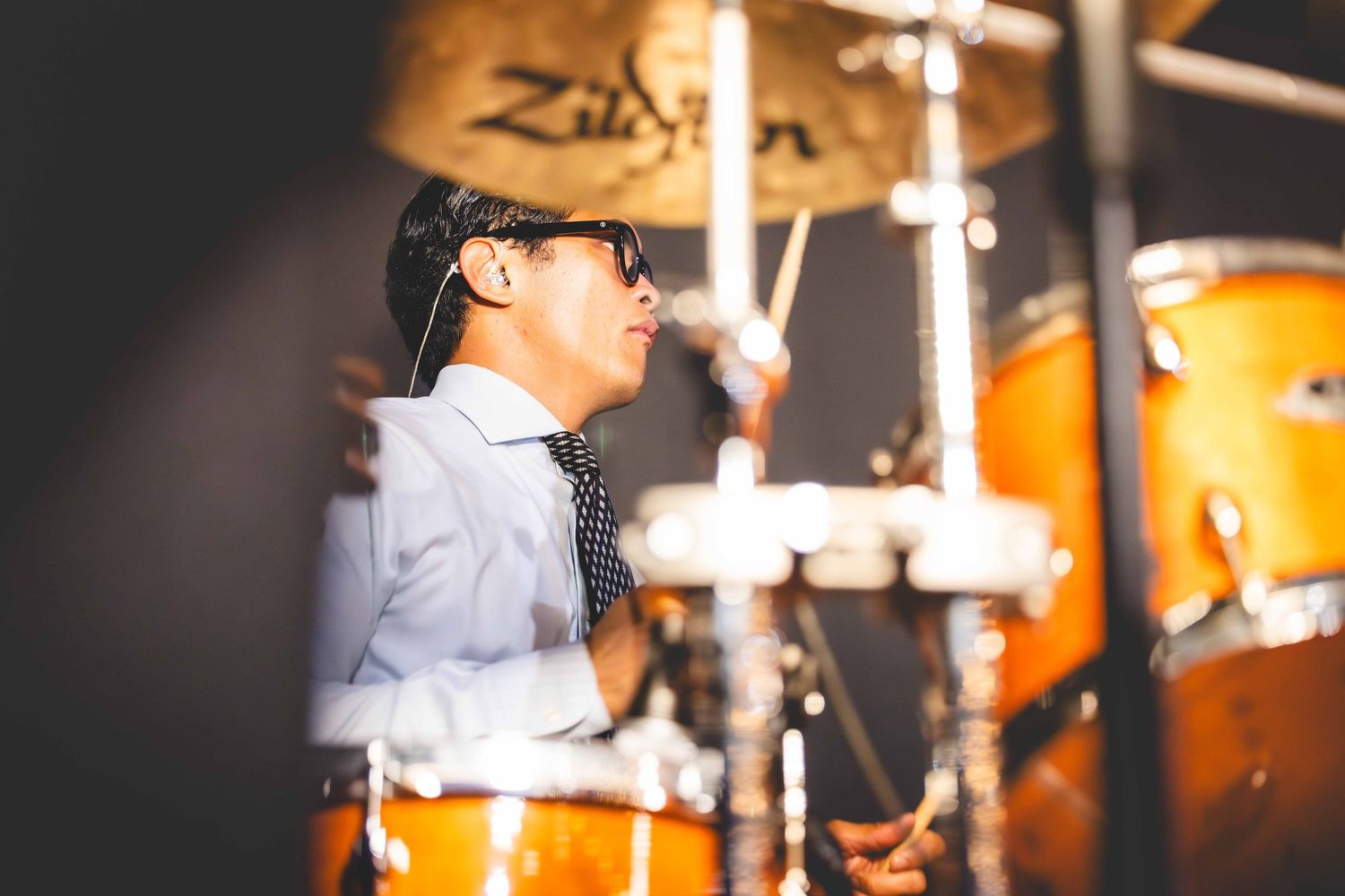 Drummer in a light blue shirt, tie and sunglasses plays an orange drum kit beneath a Zildjian cymbal during a live performance.