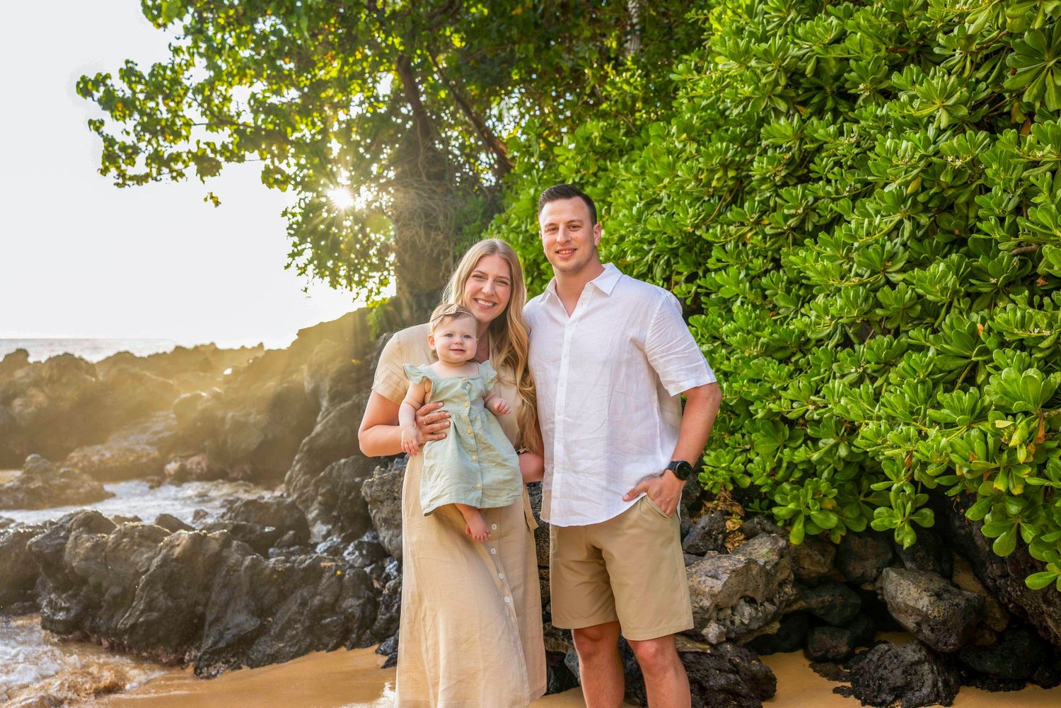 Smiling couple holding their baby girl on a rocky beach at sunset, framed by lush green foliage for a relaxed family portrait. Makena