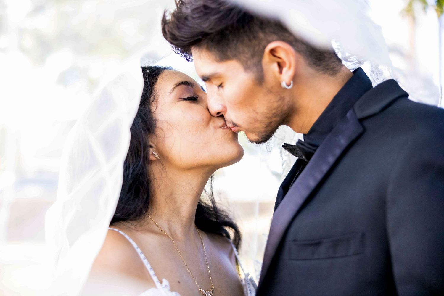 Bride and groom share a tender kiss under a sheer white veil, he in a black suit and bow tie, she in a delicate lace gown. Lahaina