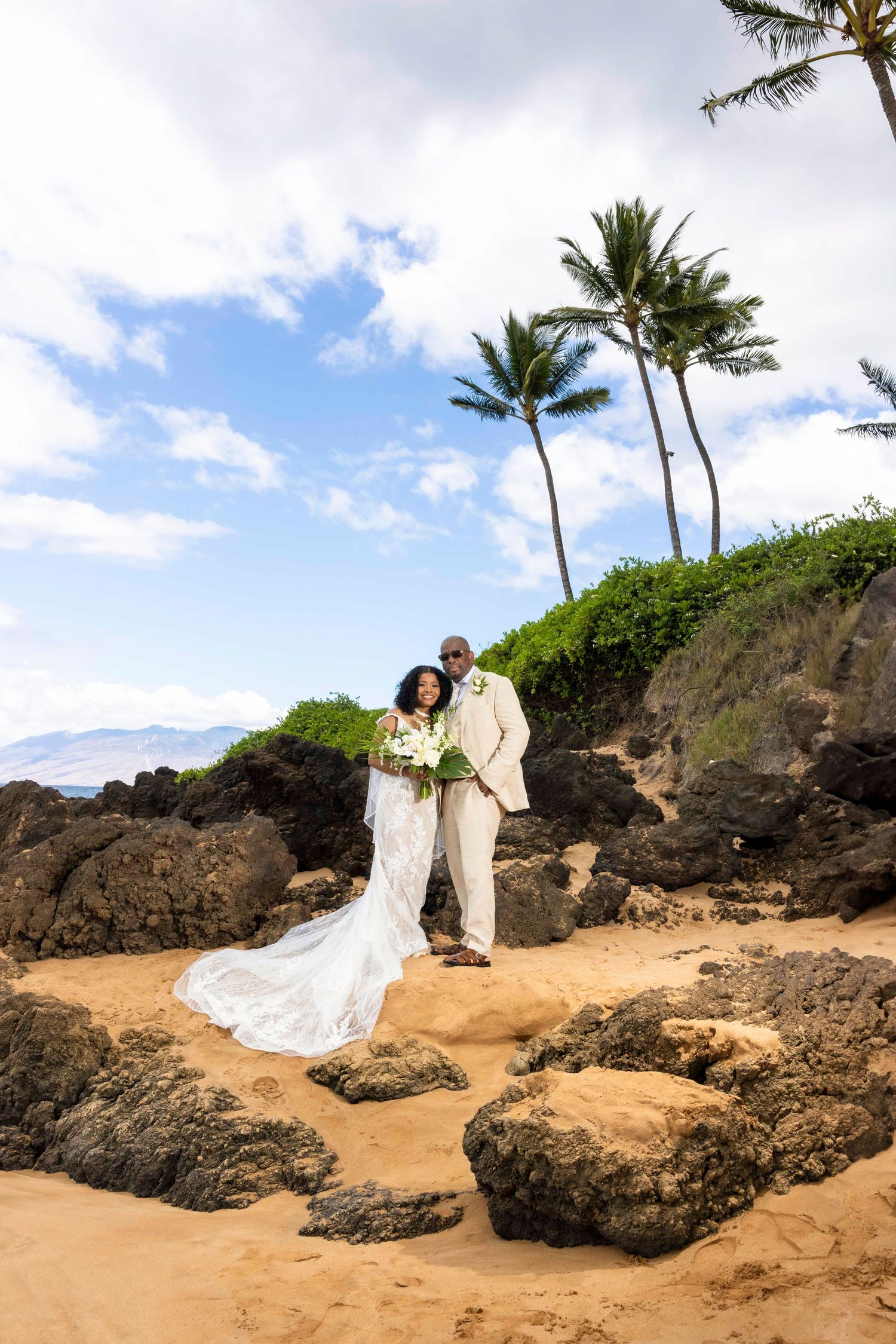 Bride in a white gown and groom in a cream suit embrace on a rocky tropical beach with palm trees and a blue sky at a destination wedding.