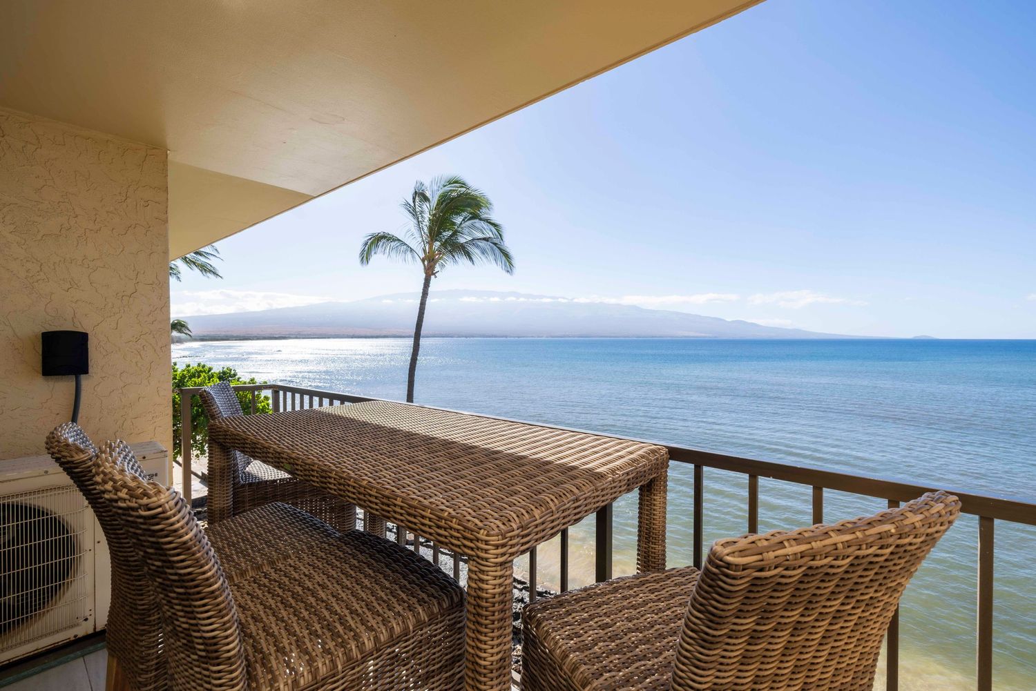 Oceanfront balcony with wicker chairs and a table overlooking a palm tree, calm blue sea, and distant mountains on a sunny tropical day.