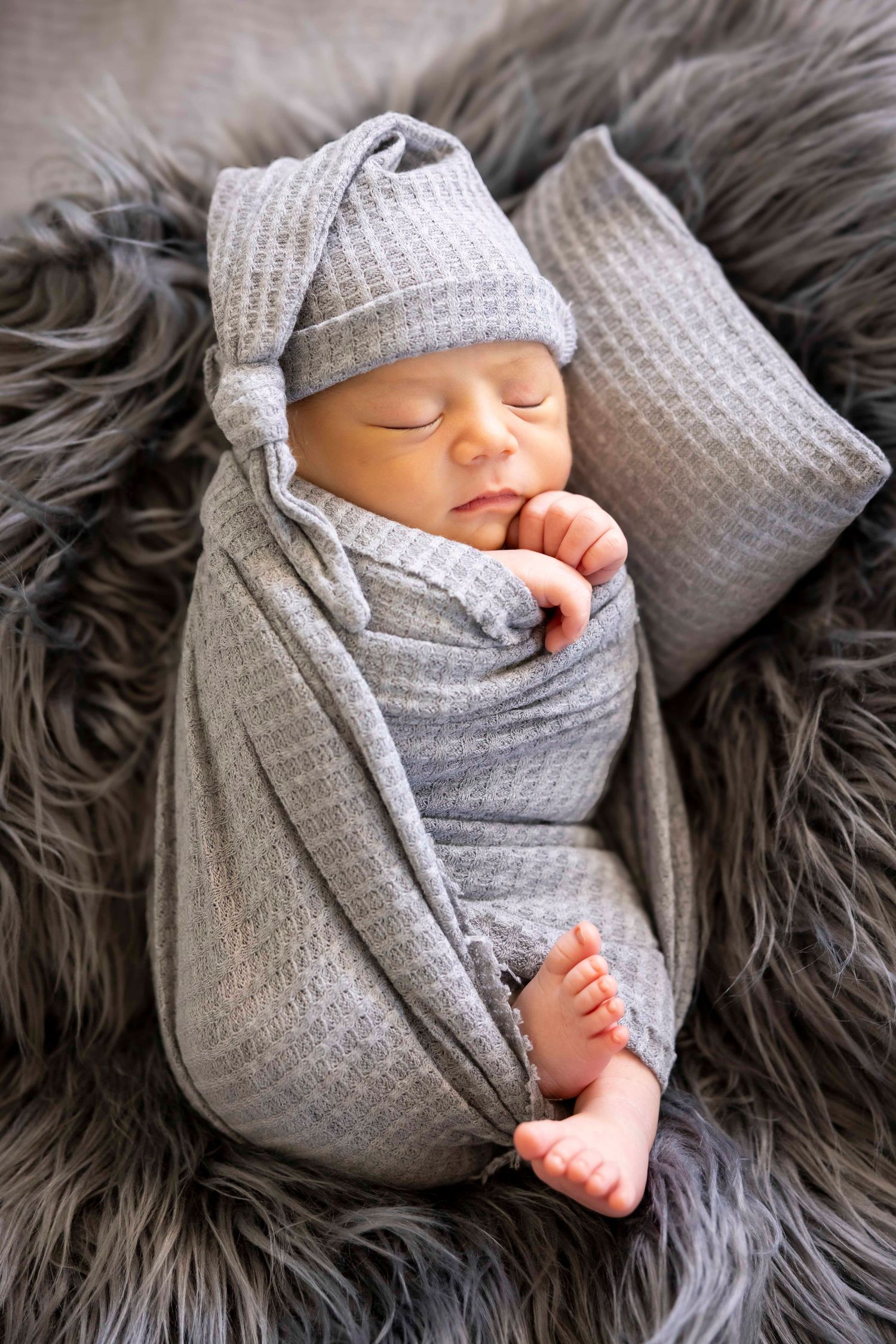 Sleeping newborn in a gray knit swaddle and knotted hat resting on a fluffy gray fur rug with a matching pillow.